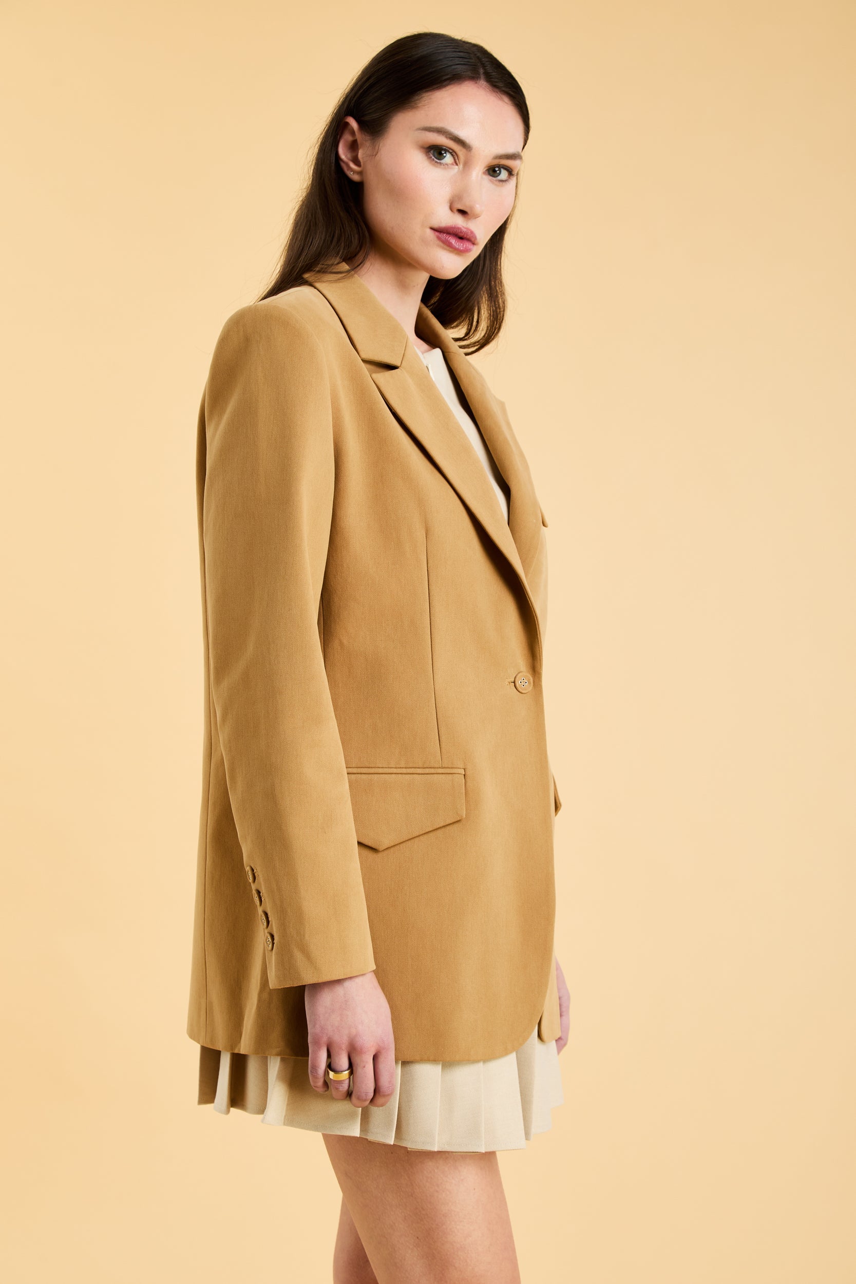 woman wearing a Camel colored Constance blazer on top of a Oat colored Elisabeth pleated mini skirt on a beige background