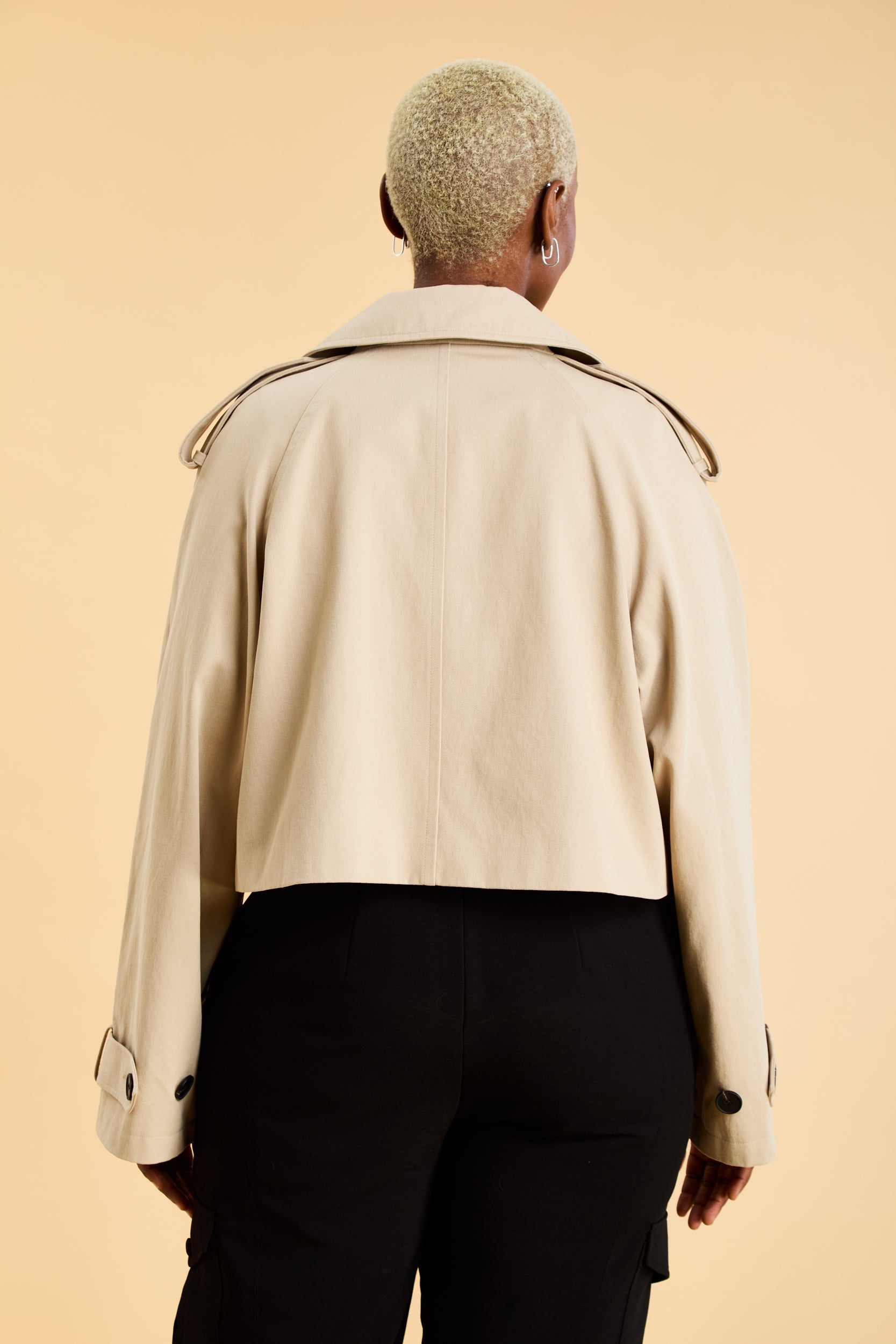 Woman wearing a beige cropped trench coat jacket in stone color against a beige background