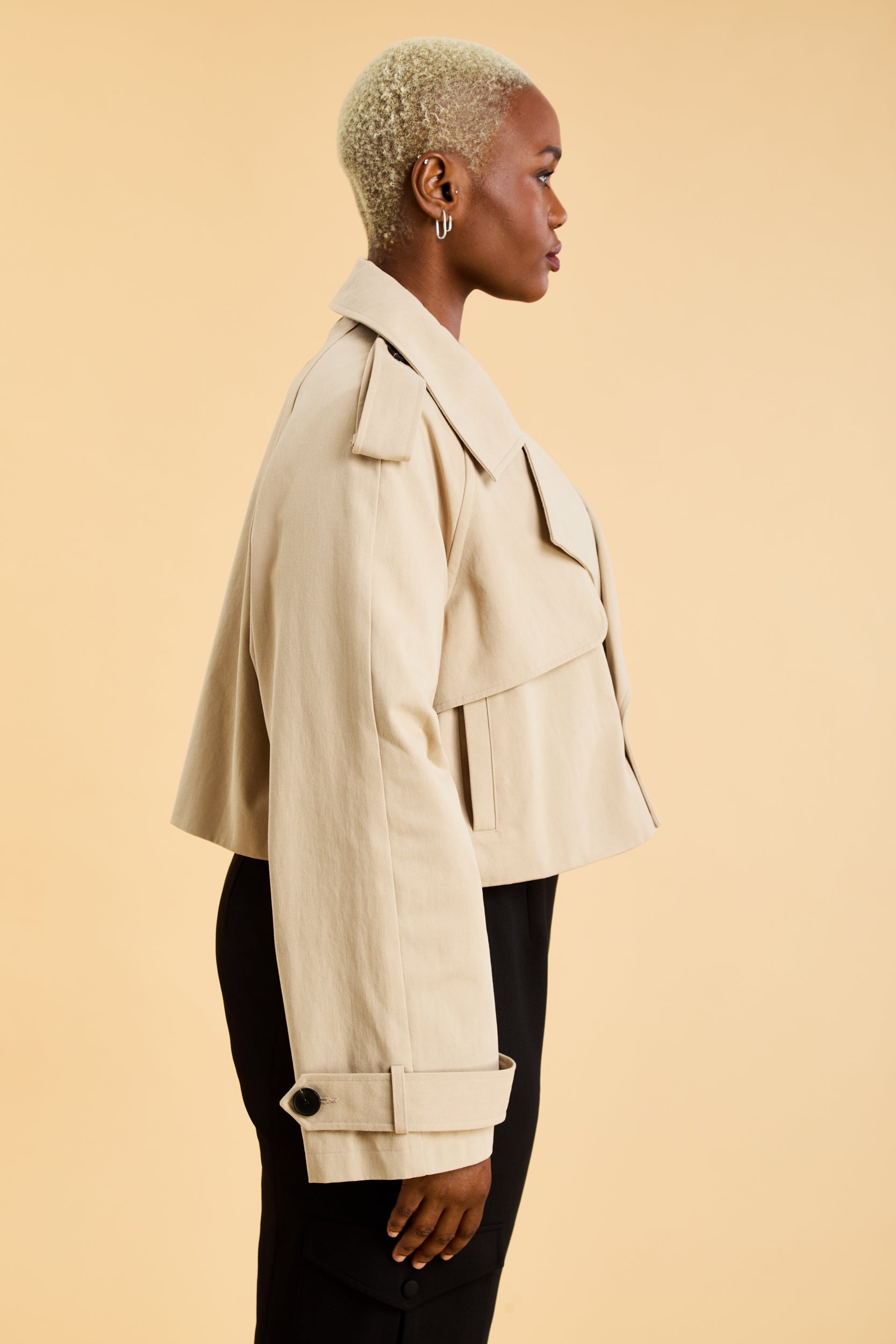 Woman wearing a beige cropped trench coat jacket in stone color against a beige background