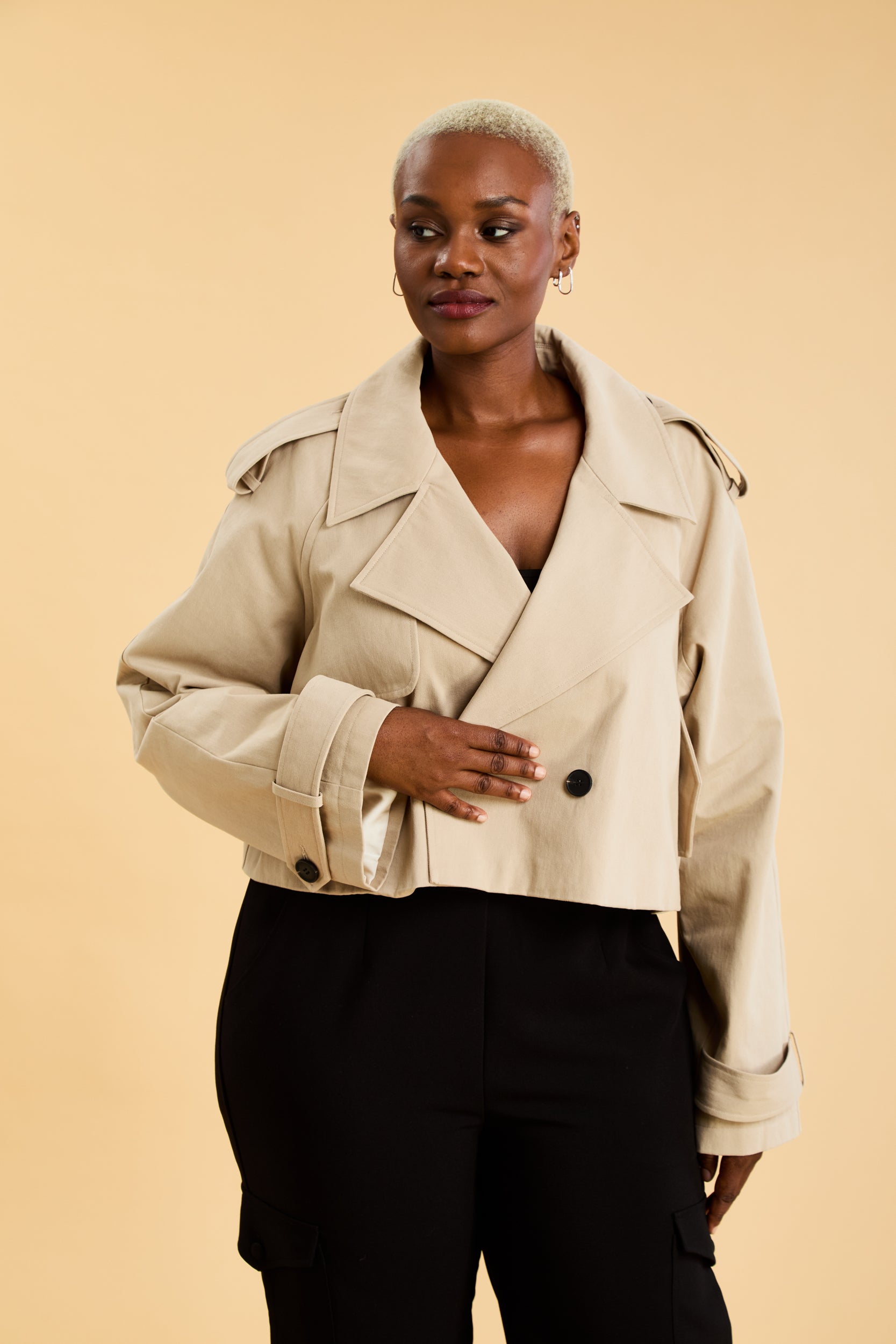 Woman wearing a beige cropped trench coat jacket in stone color against a beige background