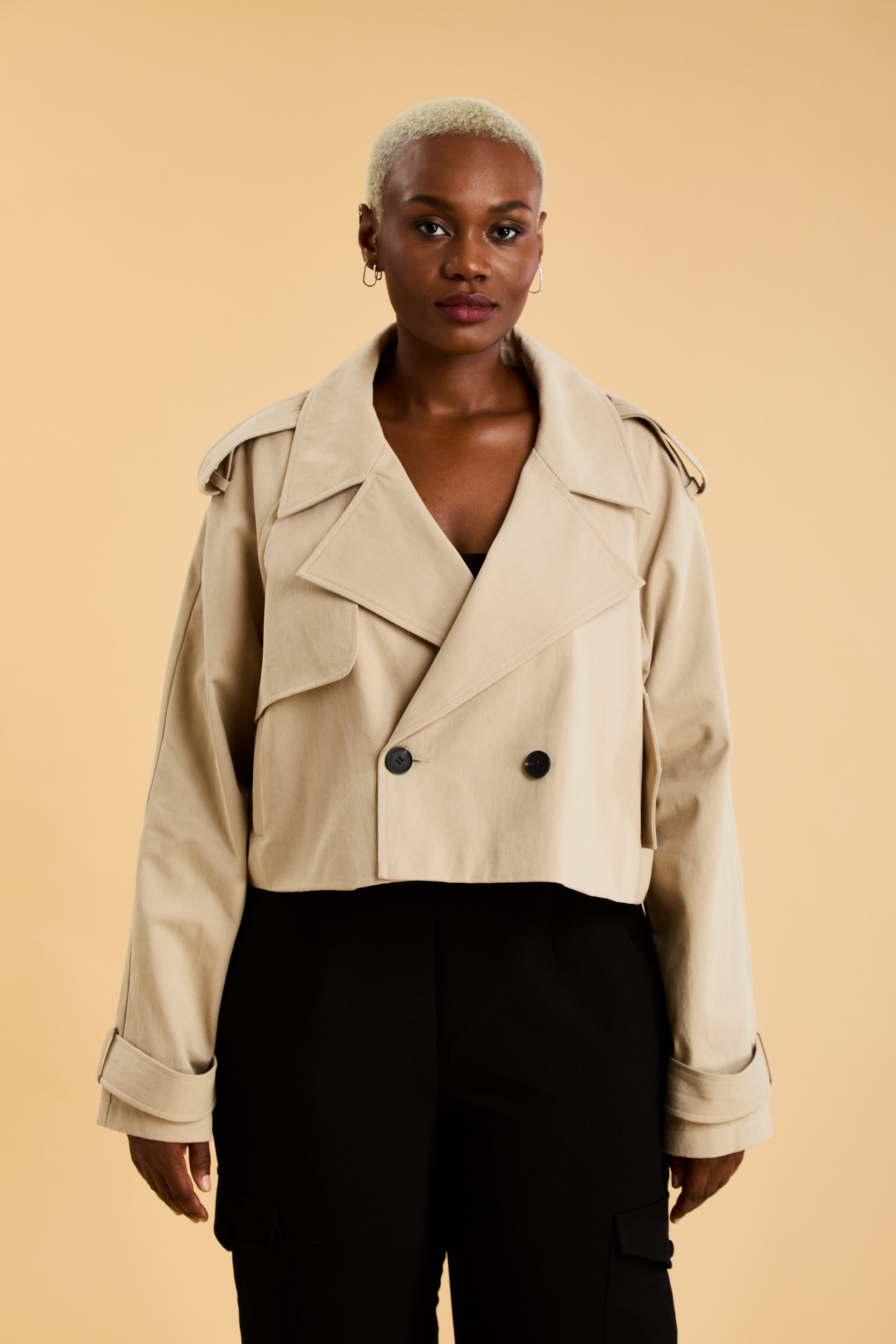 Woman wearing a beige cropped trench coat jacket in stone color against a beige background