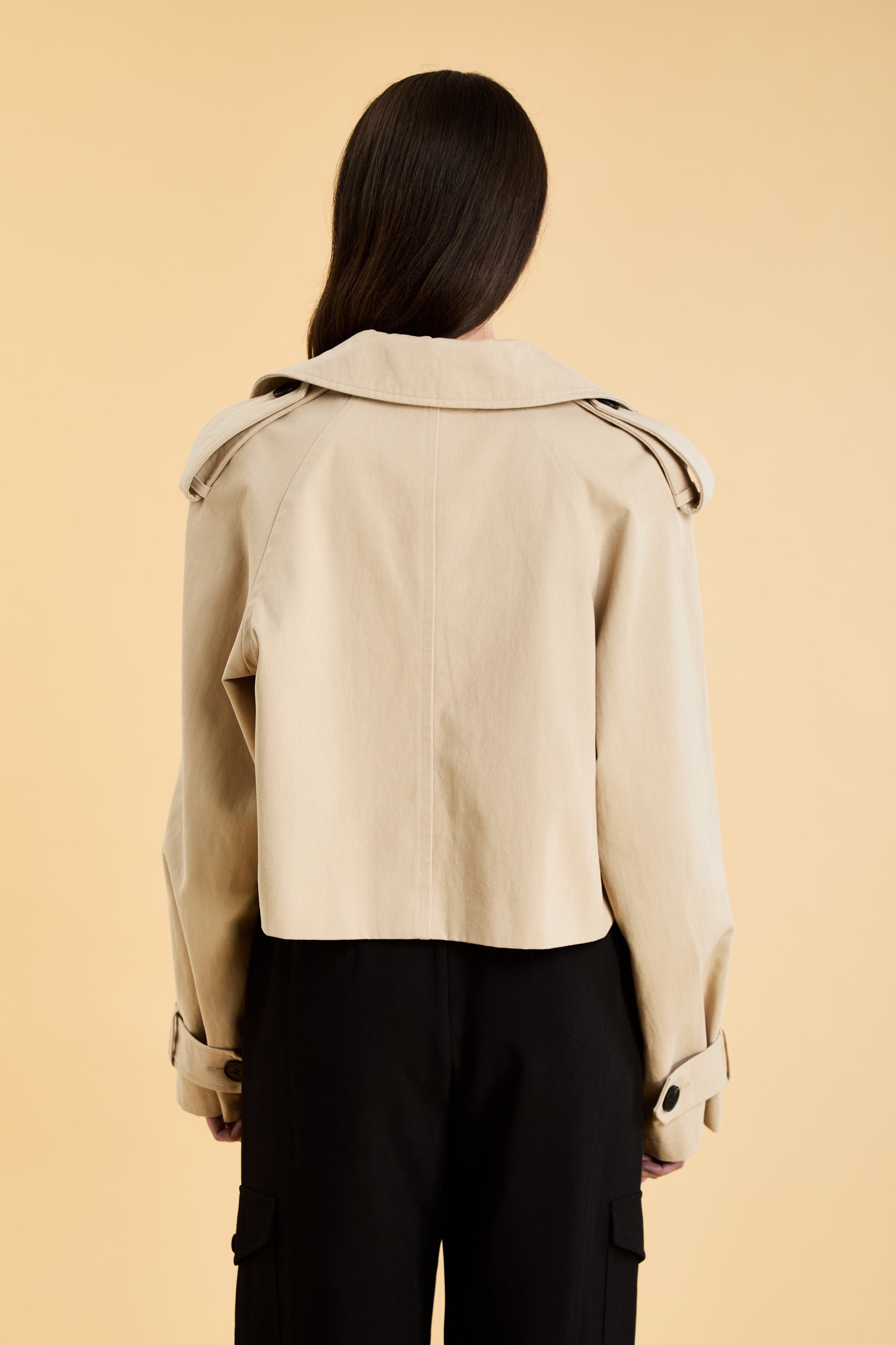 Woman wearing a beige cropped trench coat jacket in stone color against a beige background