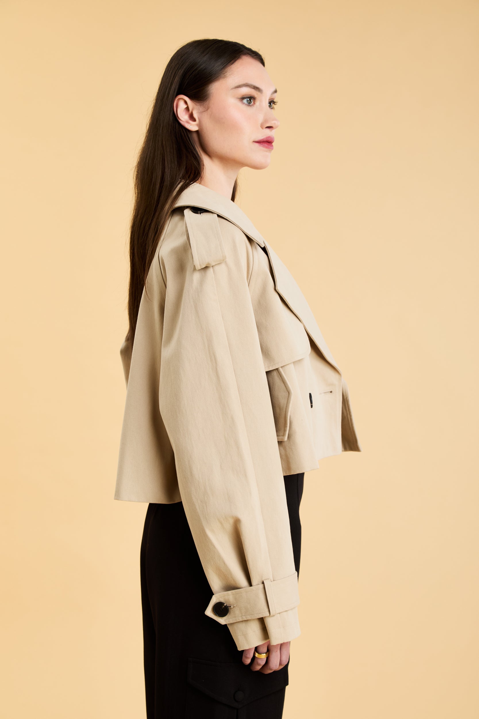 Woman wearing a beige cropped trench coat jacket in stone color against a beige background