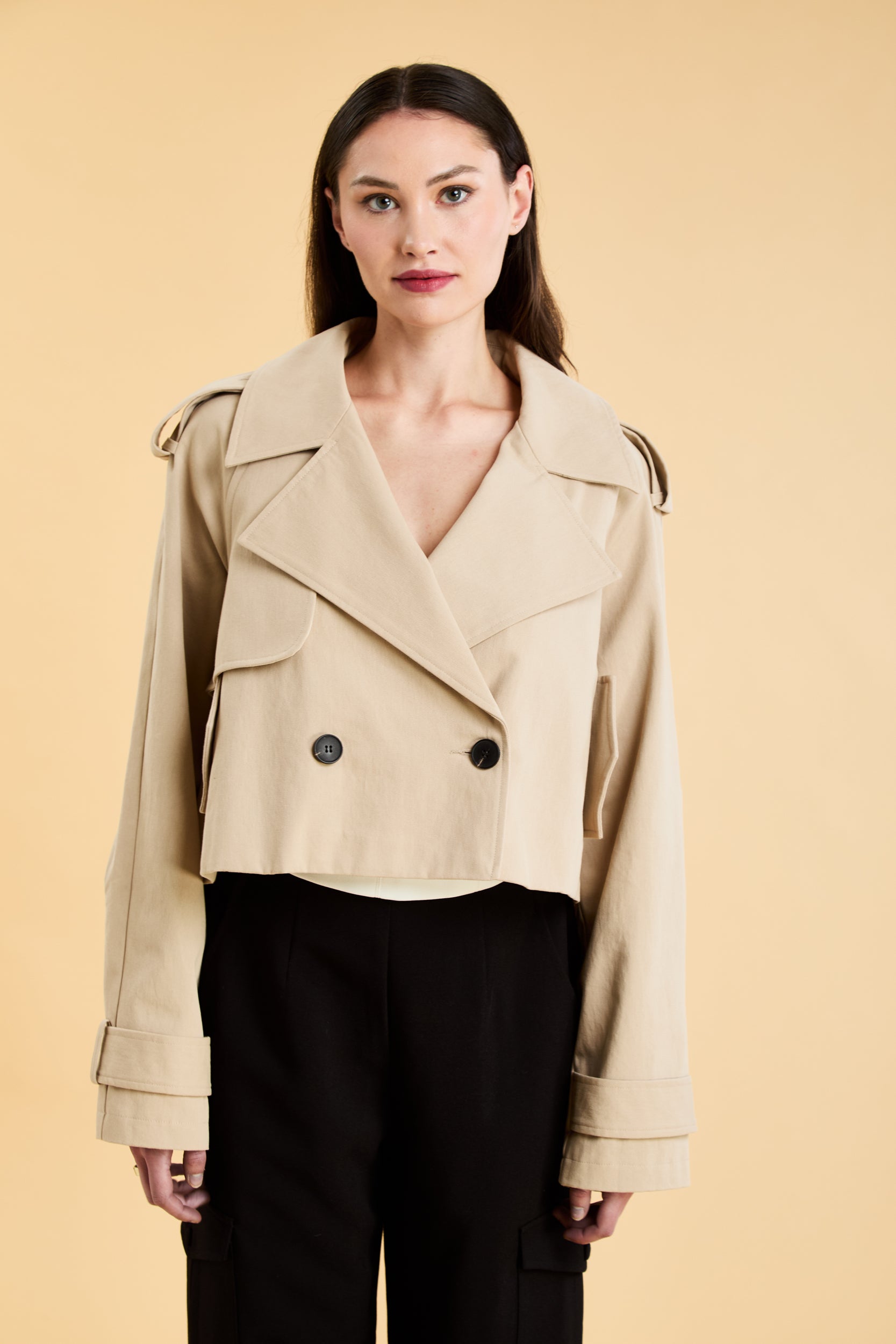 Woman wearing a beige cropped trench coat jacket in stone color against a beige background