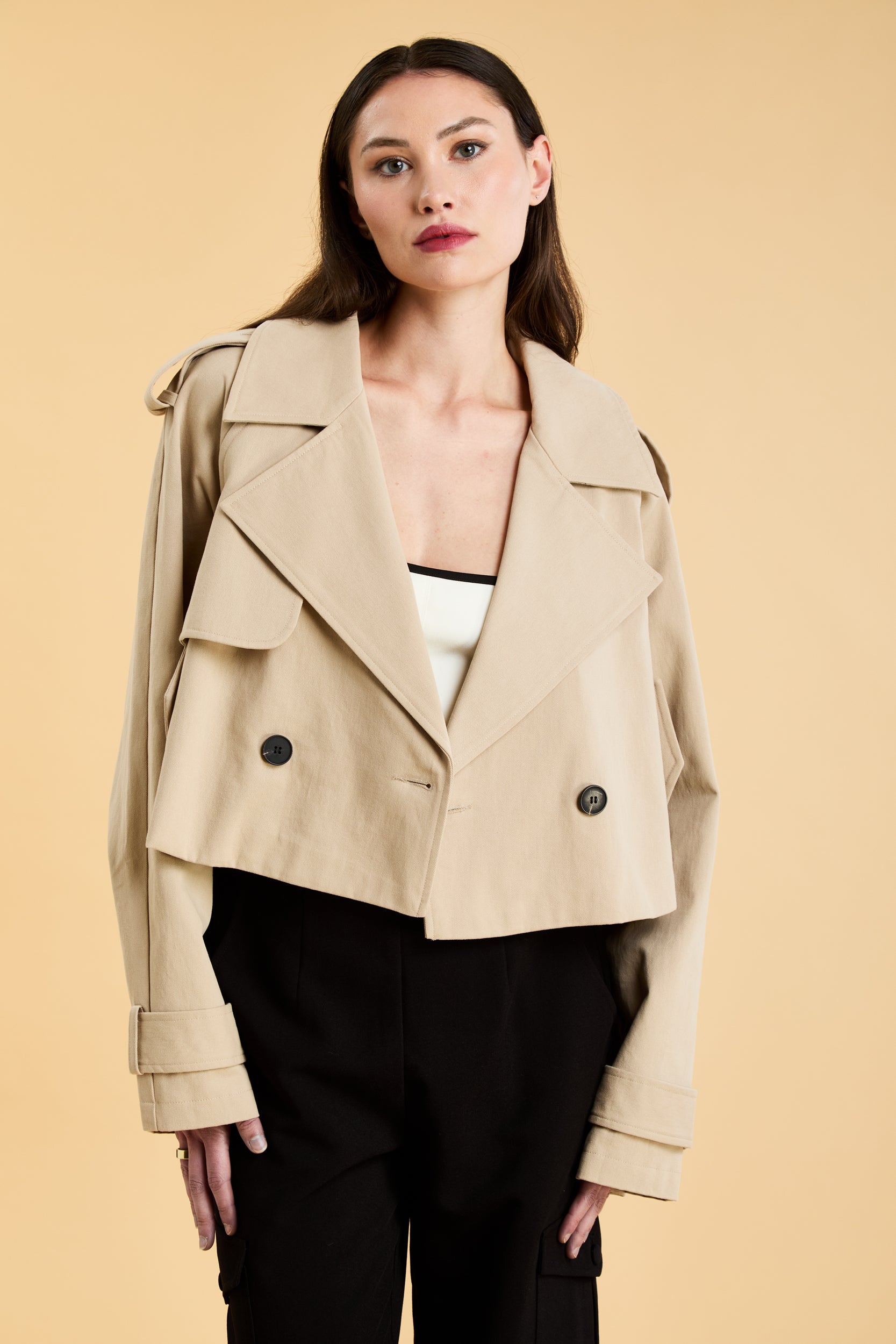 Woman wearing a beige cropped trench coat jacket in stone color against a beige background