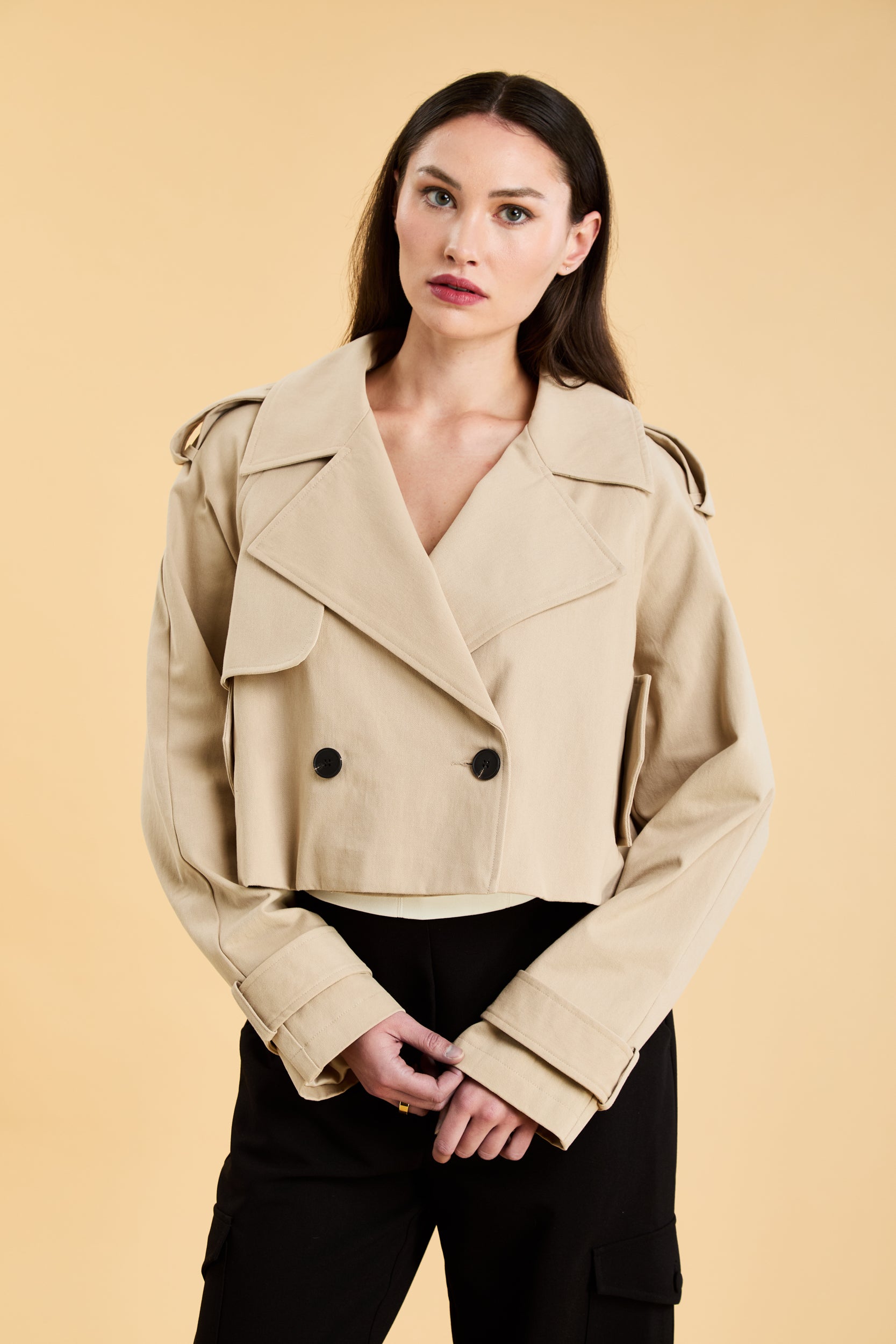 Woman wearing a beige cropped trench coat jacket in stone color against a beige background