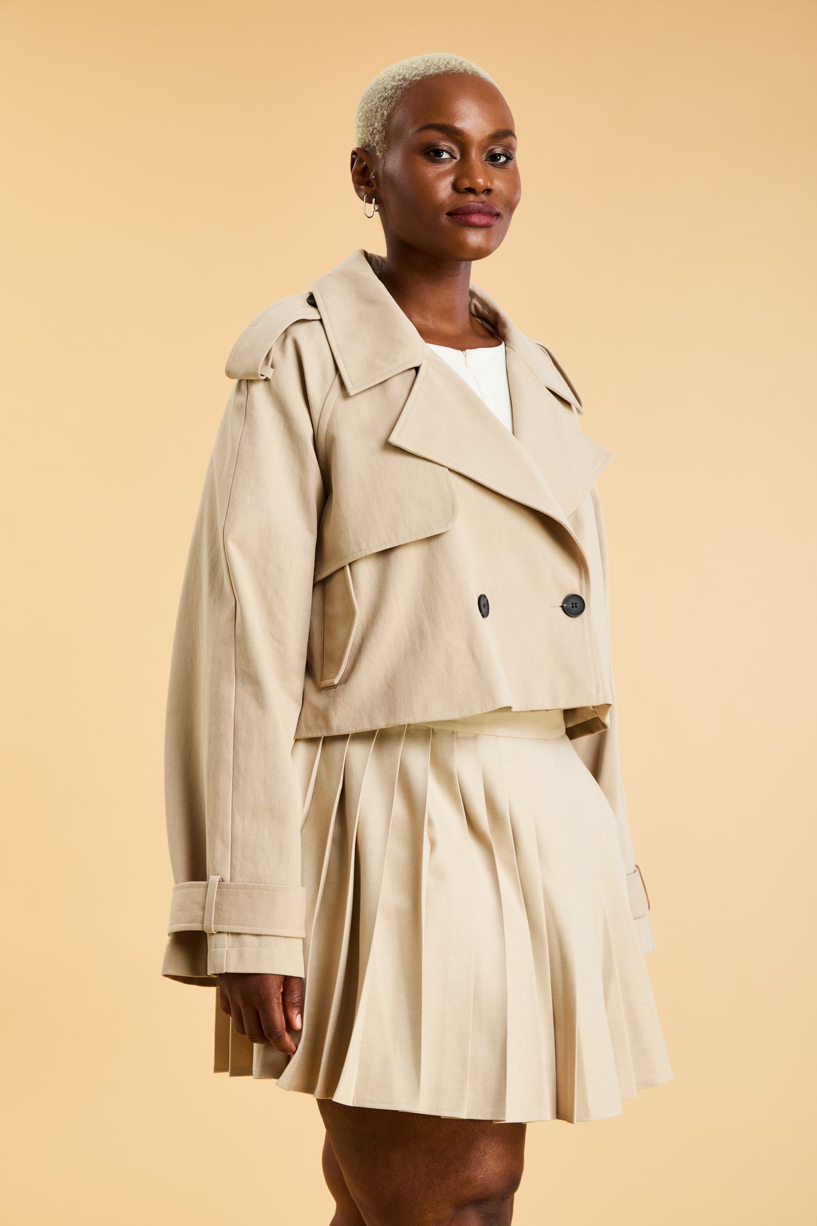 Woman wearing a beige cropped trench coat jacket in stone color against a beige background