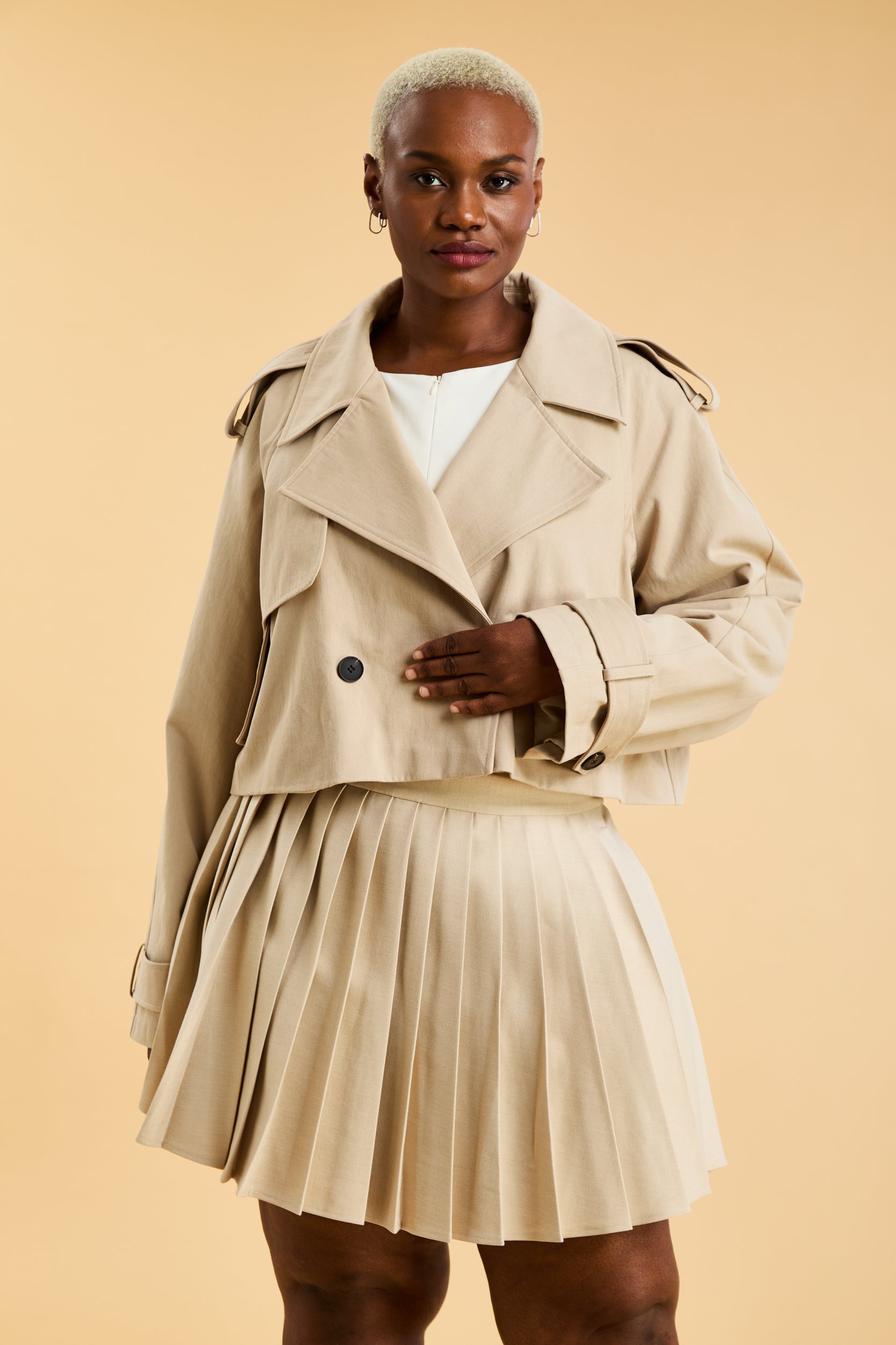Woman wearing a beige cropped trench coat jacket in stone color against a beige background