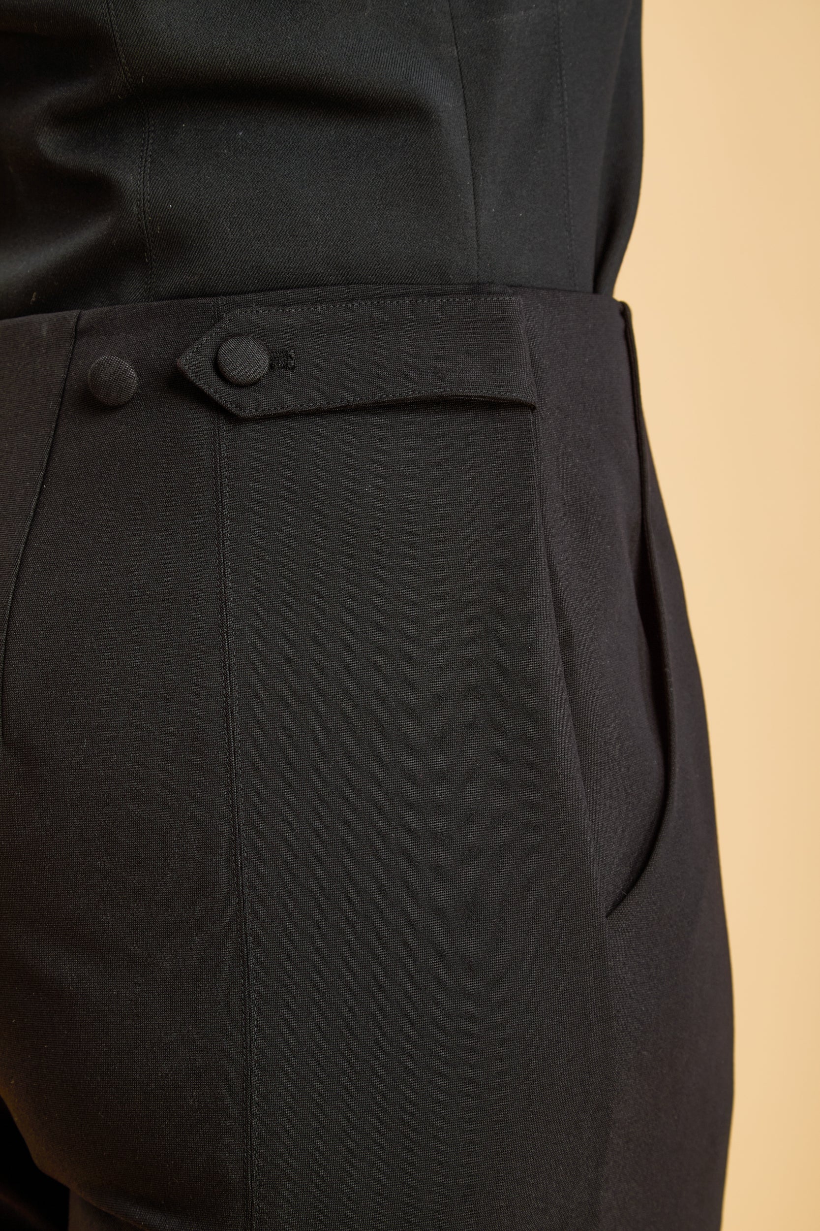 close-up of black pants with a waist tab and two buttons on a beige background