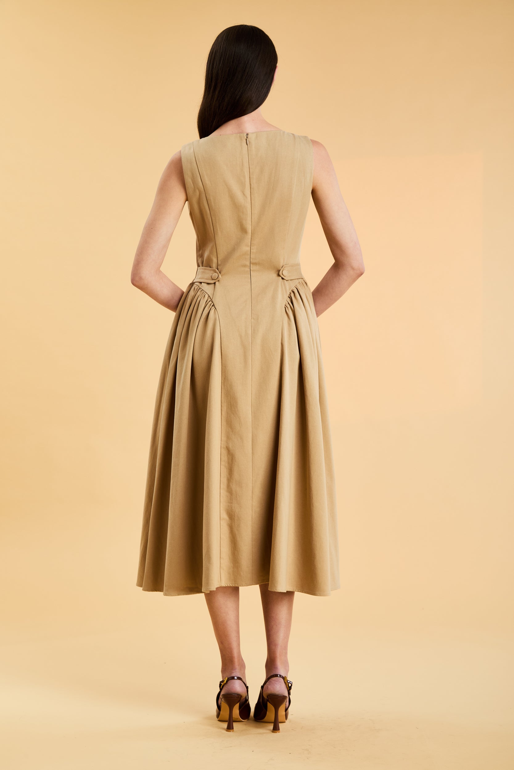 Woman wearing a Triplicity Camel colored sleeveless midi dress with gathered side panels on a beige background