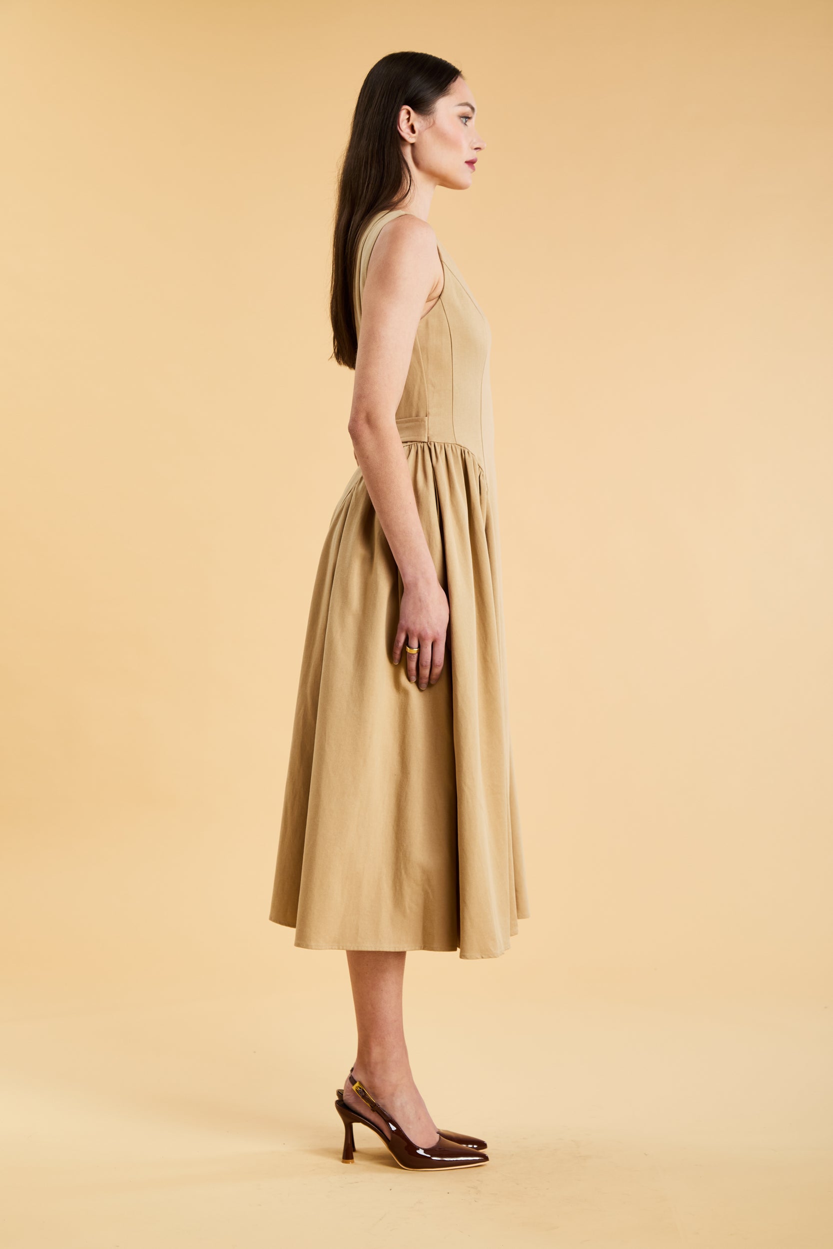 Woman wearing a Triplicity Camel colored sleeveless midi dress with gathered side panels on a beige background