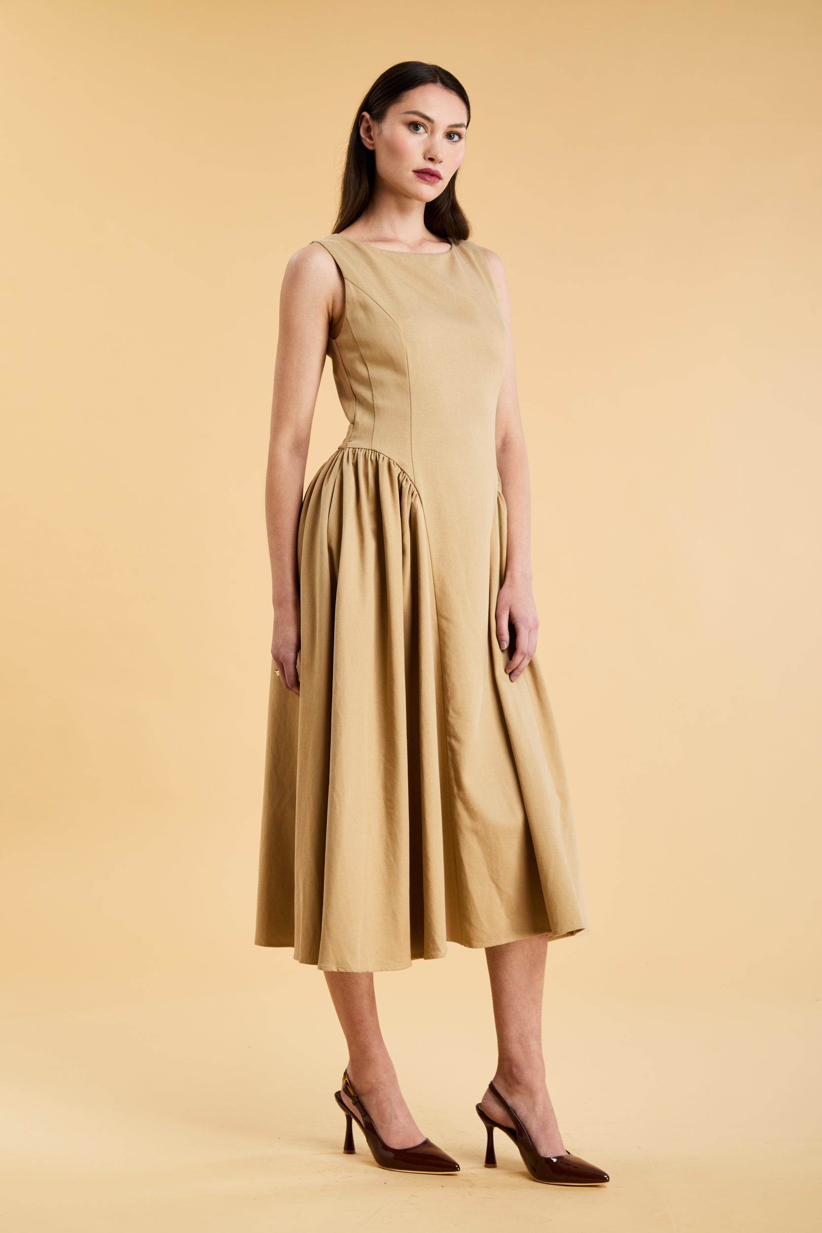 Woman wearing a Triplicity Camel colored sleeveless midi dress with gathered side panels on a beige background