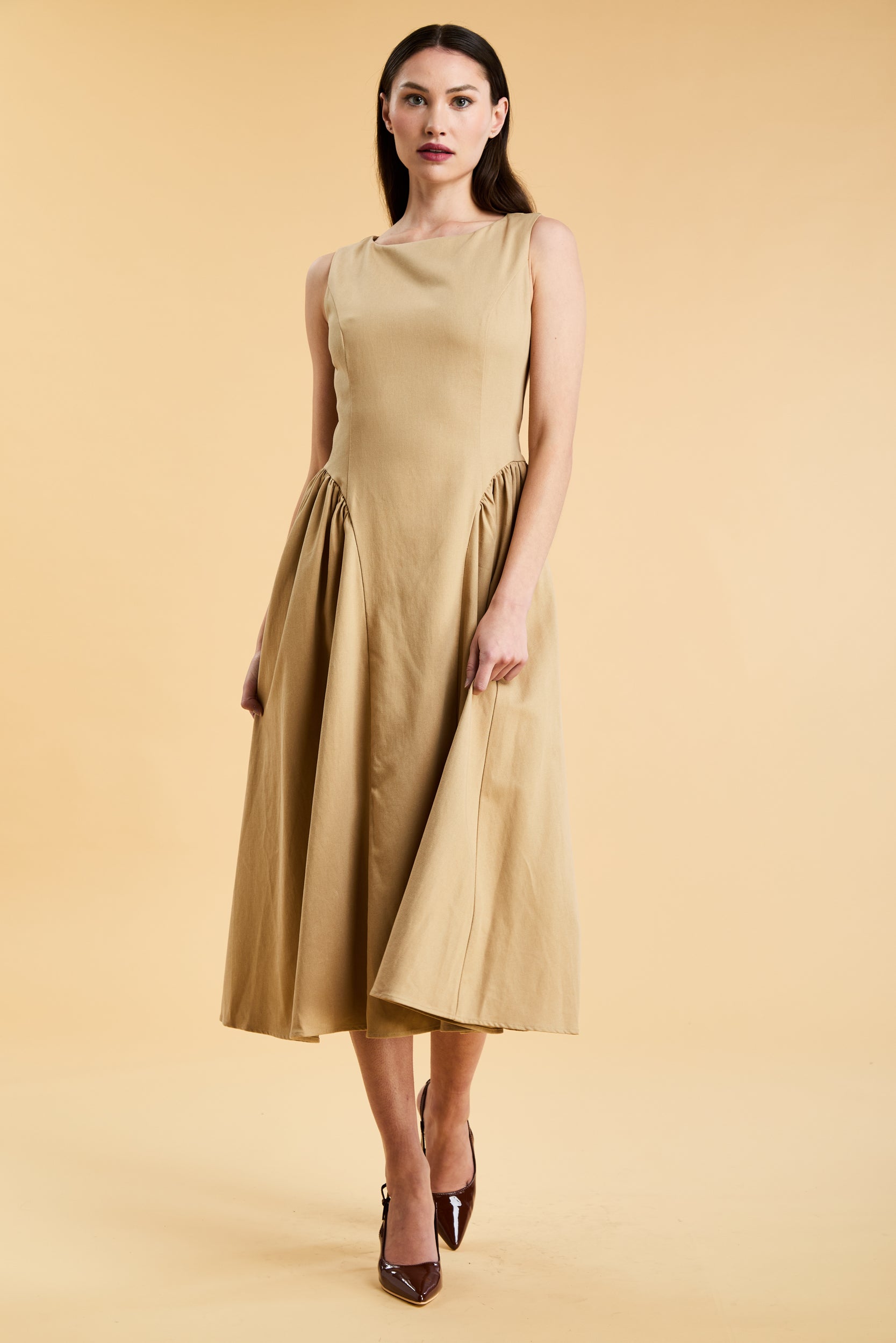 Woman wearing a Triplicity Camel colored sleeveless midi dress with gathered side panels on a beige background