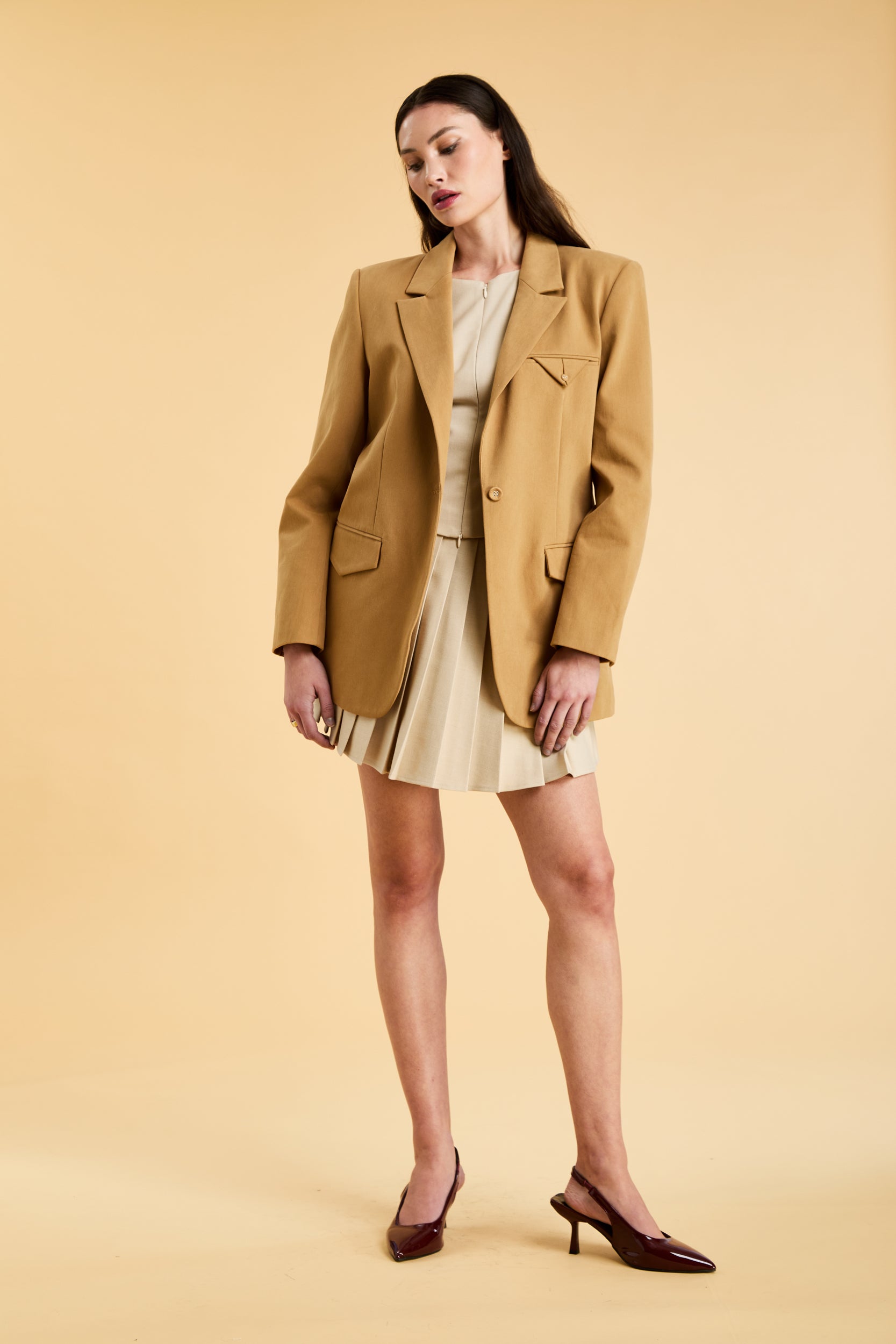 Woman wearing a Camel colored blazer and Oat colored pleated skirt on a beige background