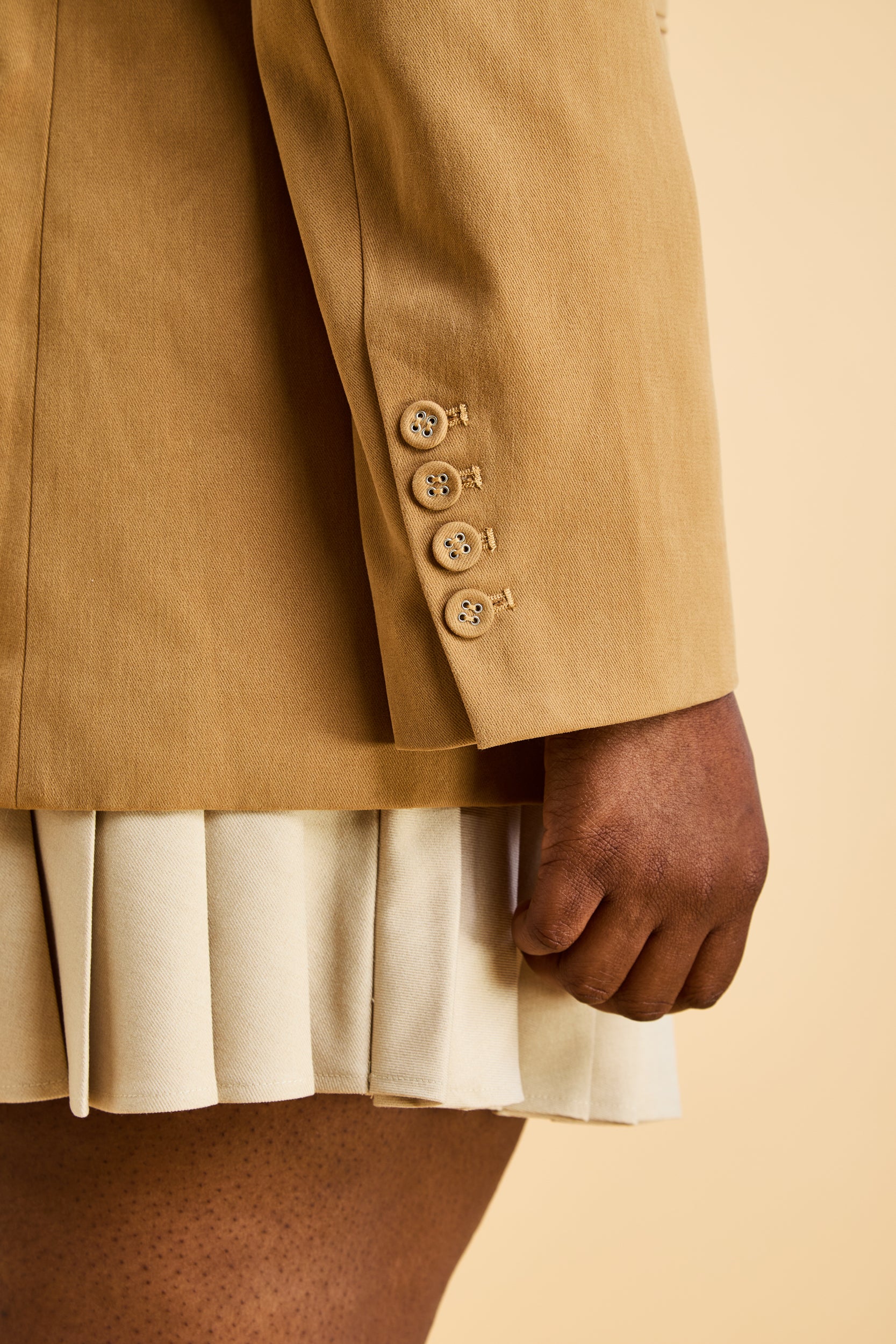 Close-up of a person wearing a Camel colored coat with buttons on the back of a detailed two piece sleeve on a beige background