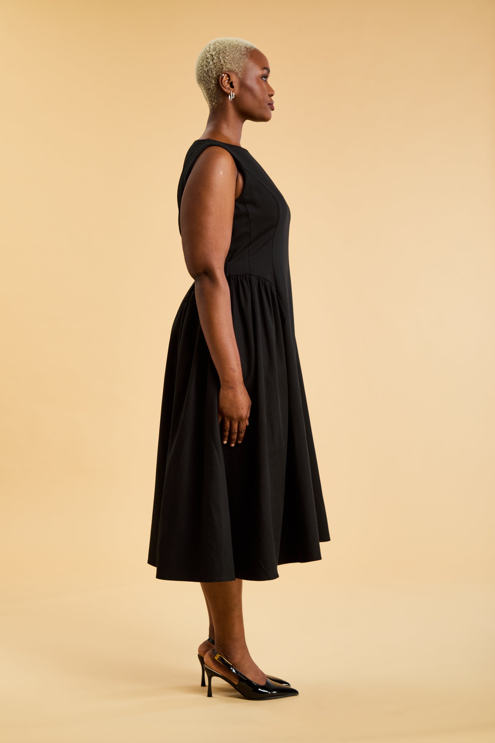 Woman wearing a Triplicity Black sleeveless midi dress with gathered side panels on a beige background