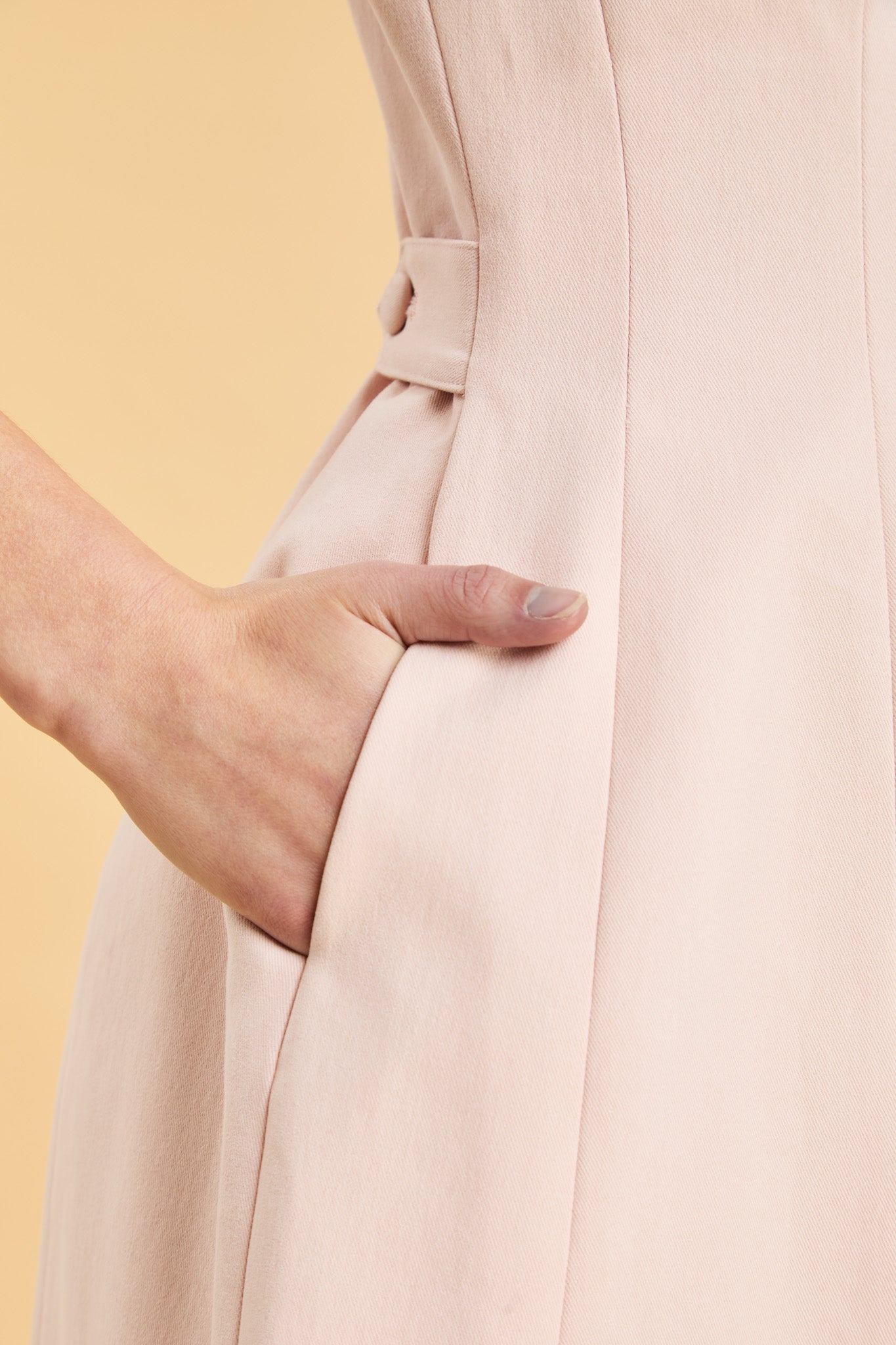 Close-up of a person wearing a blush pink dress with her hand in her pocket and a yellow background