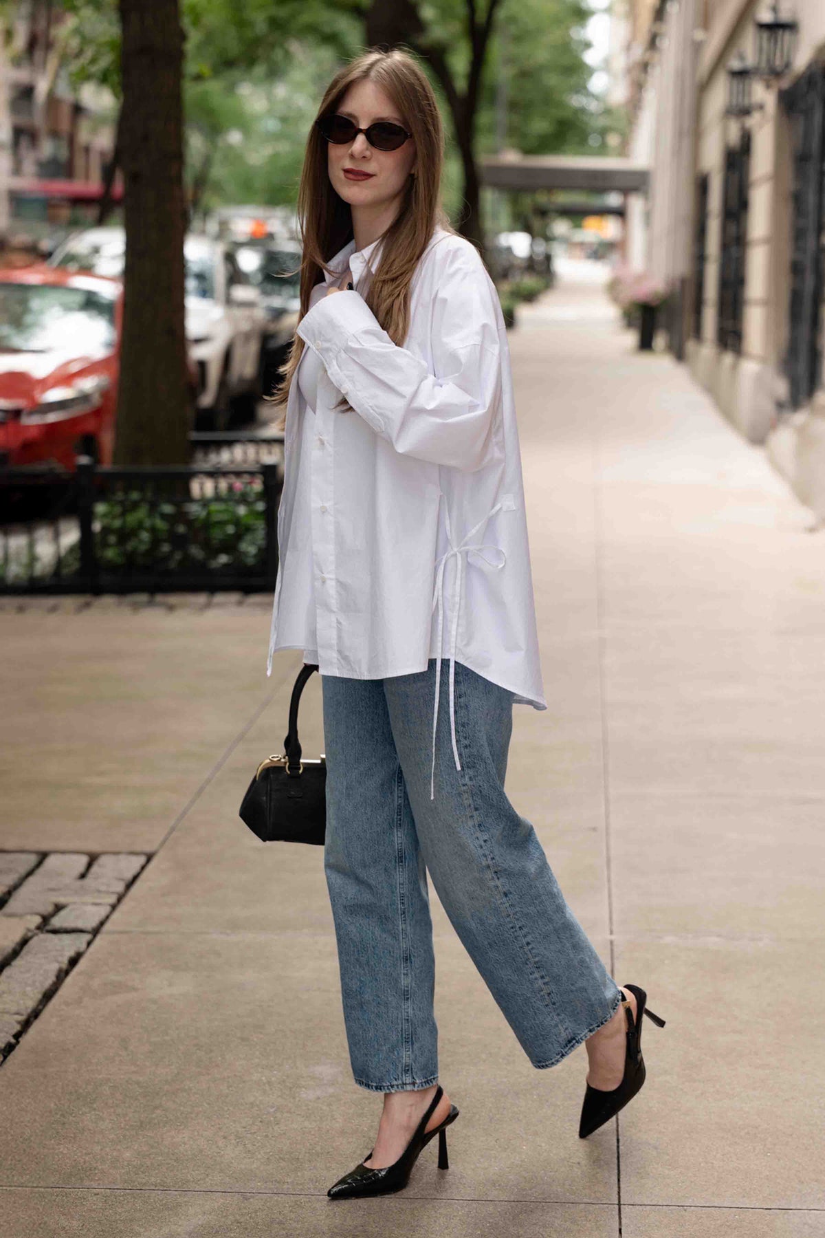 A woman walking on a city sidewalk wearing an oversized white long-sleeve shirt, wide-leg light blue jeans, black pointed-toe mule heels, and carrying a small black handbag.