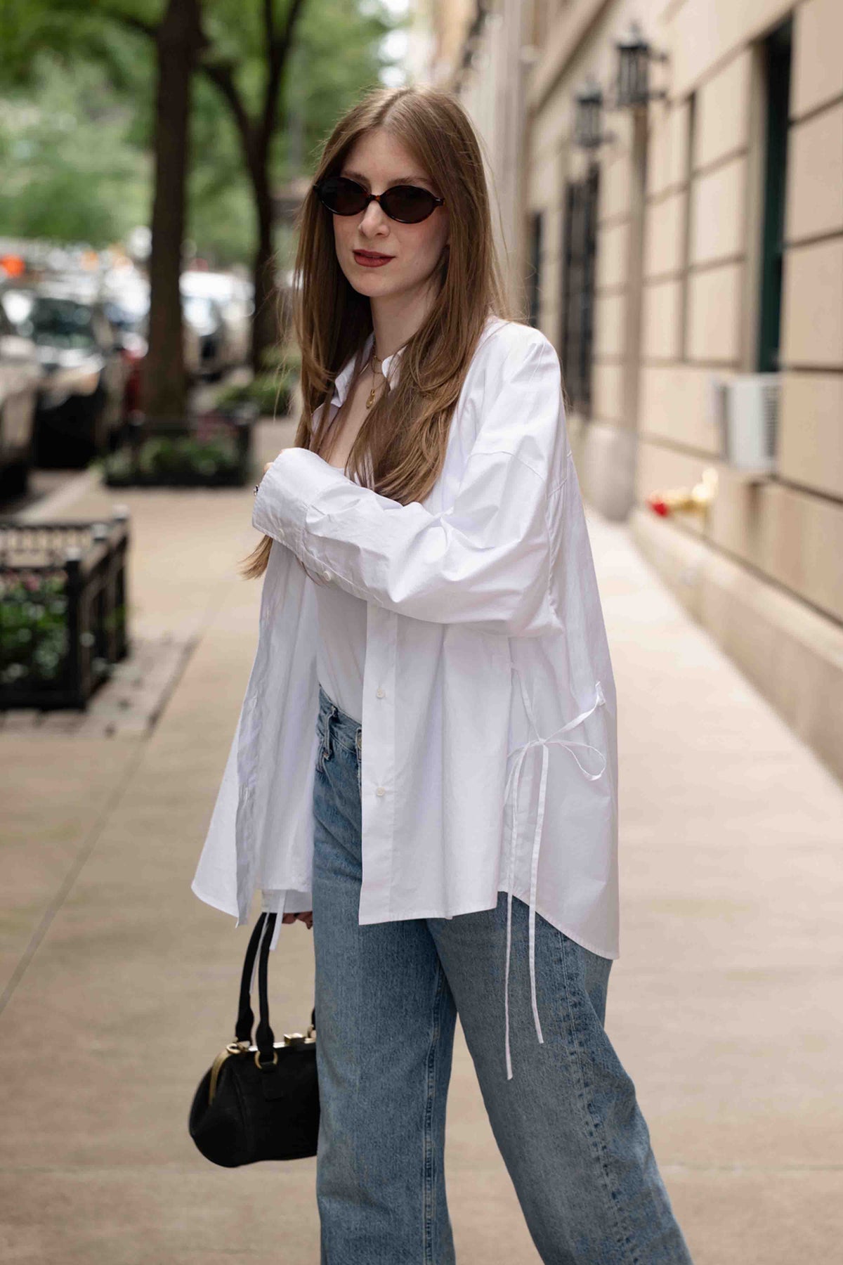 A woman stands on a city sidewalk wearing an oversized white button-up shirt, light blue jeans, dark sunglasses, and holding a small black handbag.