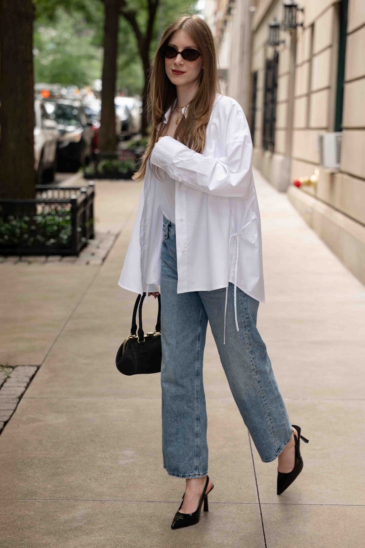 A woman walking on a city sidewalk wearing an oversized white long-sleeve blouse, light-wash wide-leg jeans, black pointed-toe heels, and carrying a small black handbag.