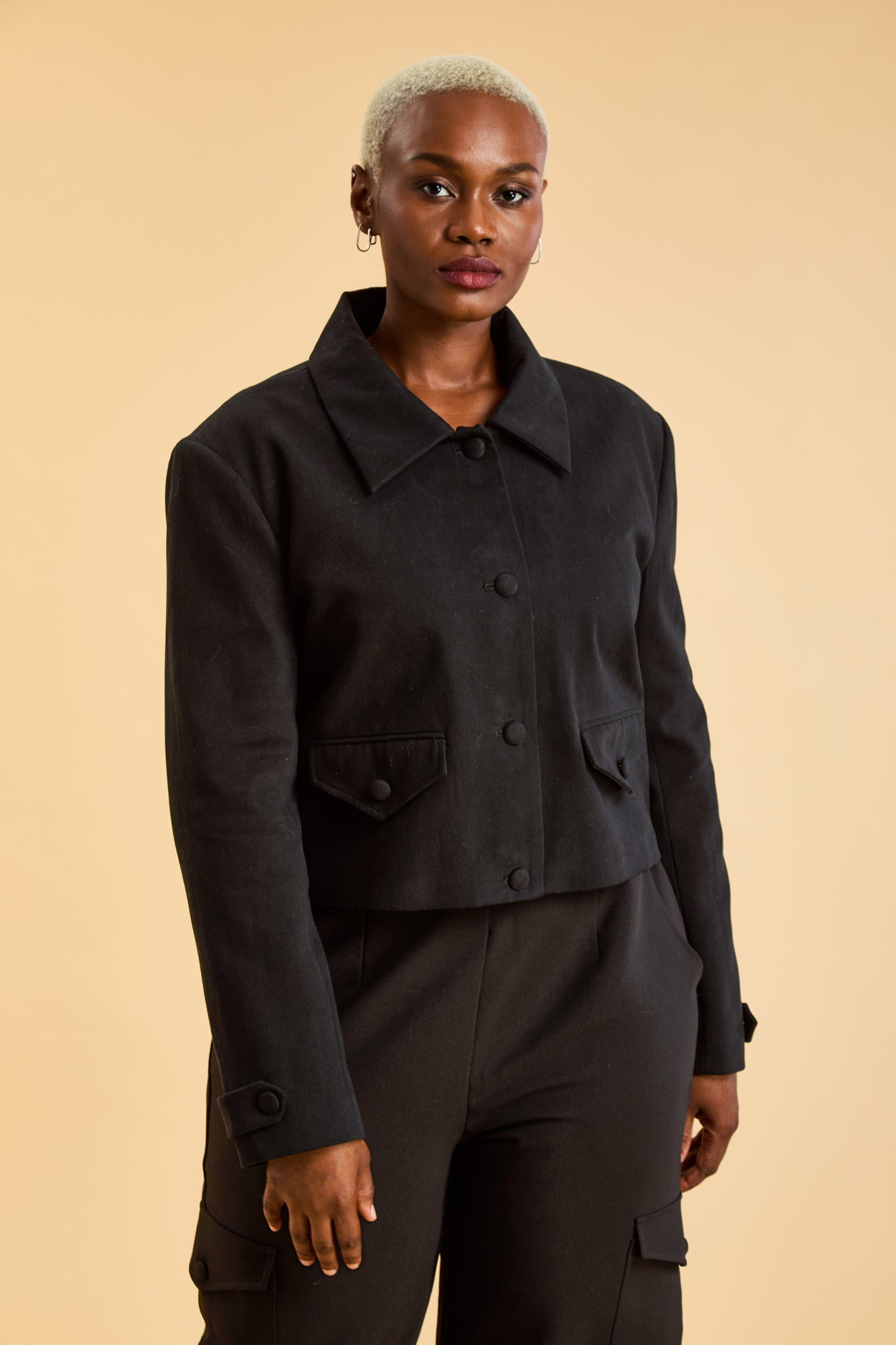 Woman wearing a Black Triplicity collared jacket and Black Triplicity pants on a beige background front close up