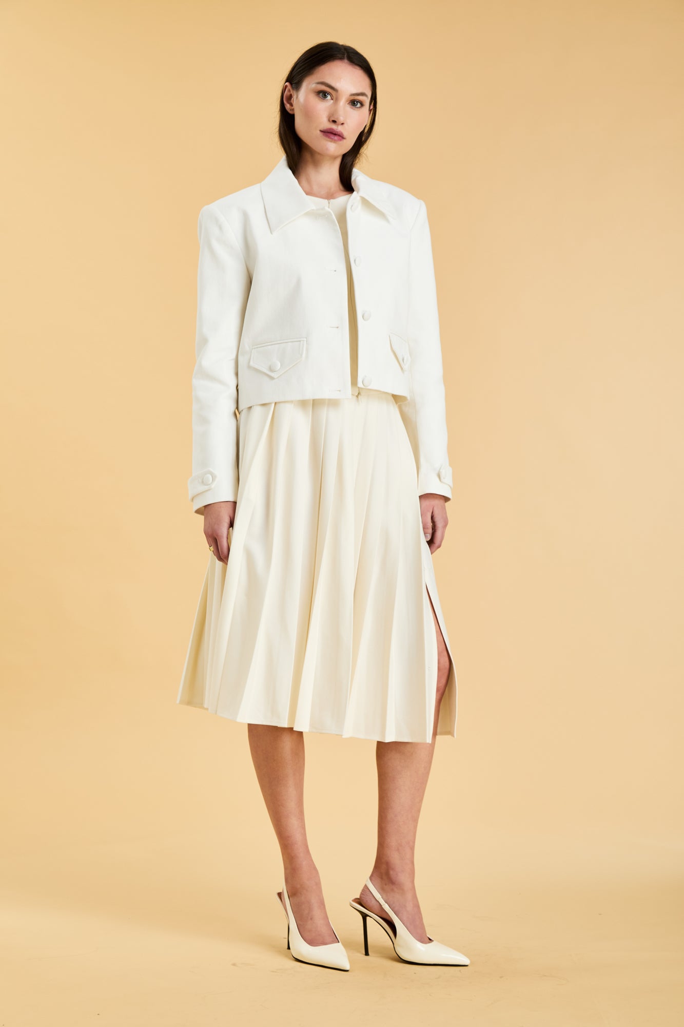 Woman wearing a White Triplicity collared tailored jacket and a White Triplicity pleated skirt against a beige background front view full body