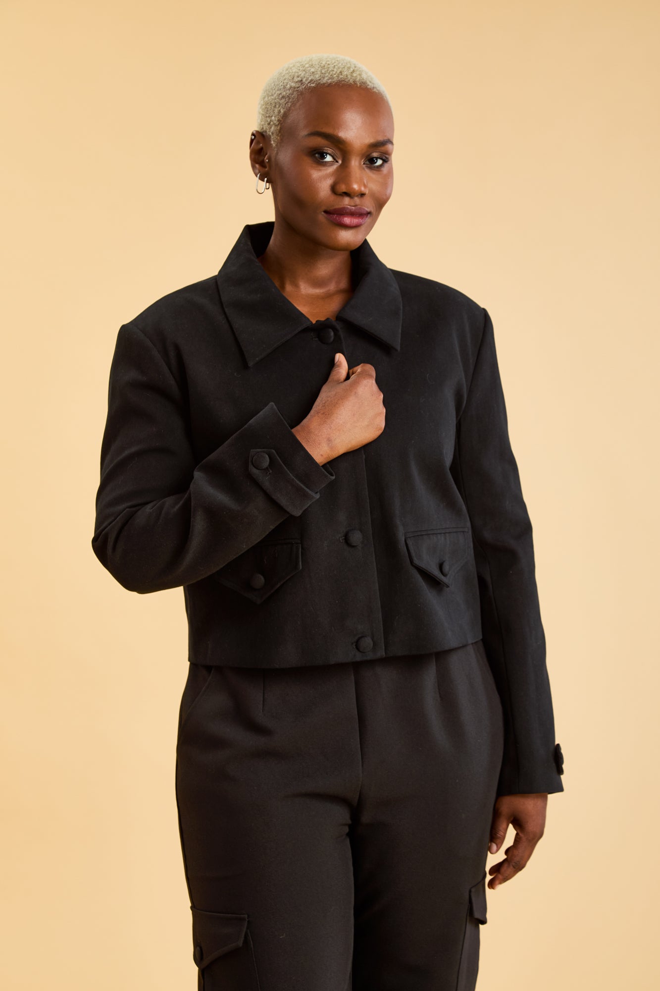 Woman wearing a Black Triplicity collared jacket and Black Triplicity pants on a beige background front close up