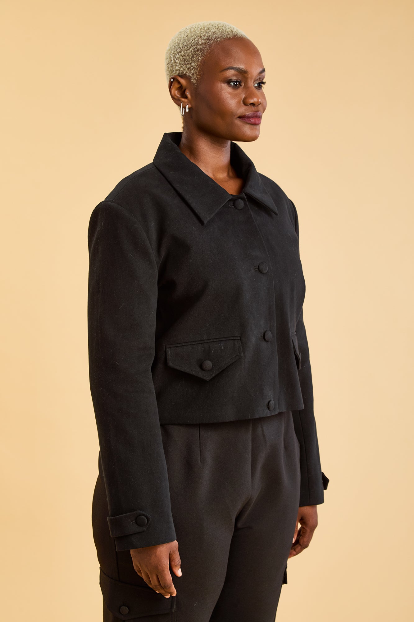 Woman wearing a Black Triplicity collared jacket and Black Triplicity pants on a beige background three quarter view close up