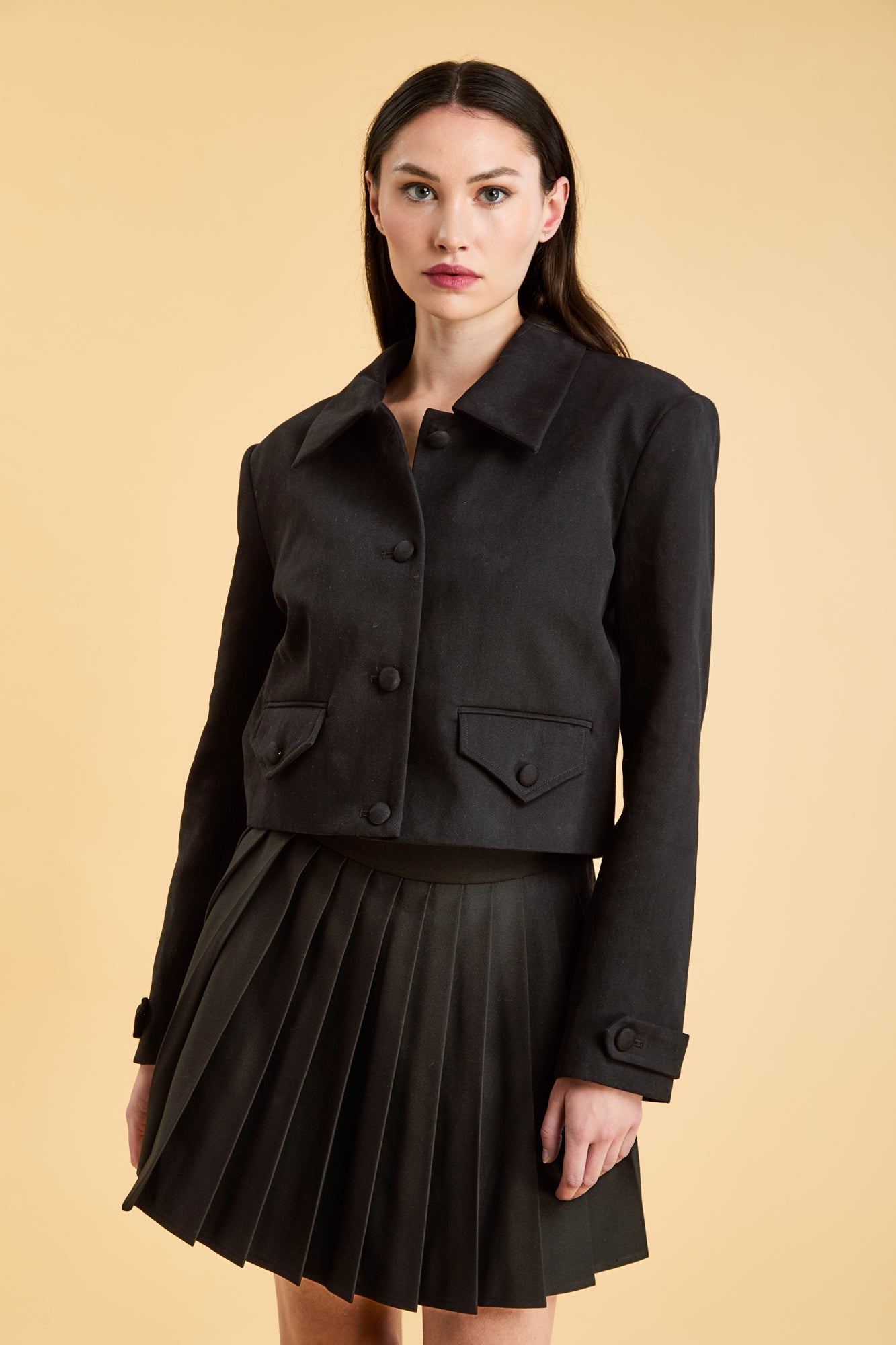 Woman wearing a Black Triplicity collared jacket and Black Triplicity pleated skirt on a beige background front close up