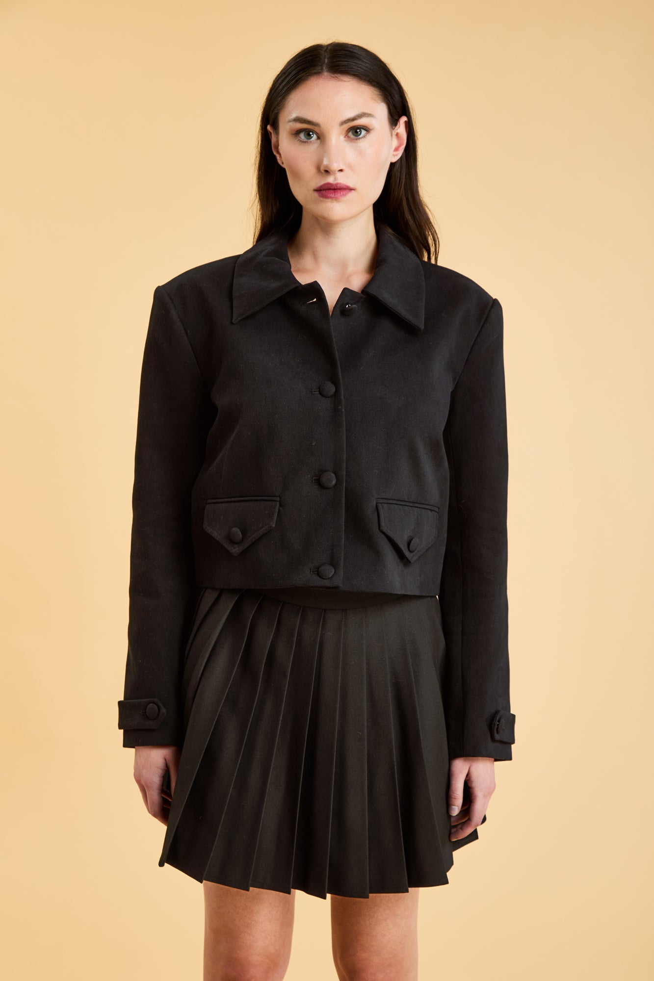 Woman wearing a Black Triplicity collared jacket and Black Triplicity pleated skirt on a beige background front close up