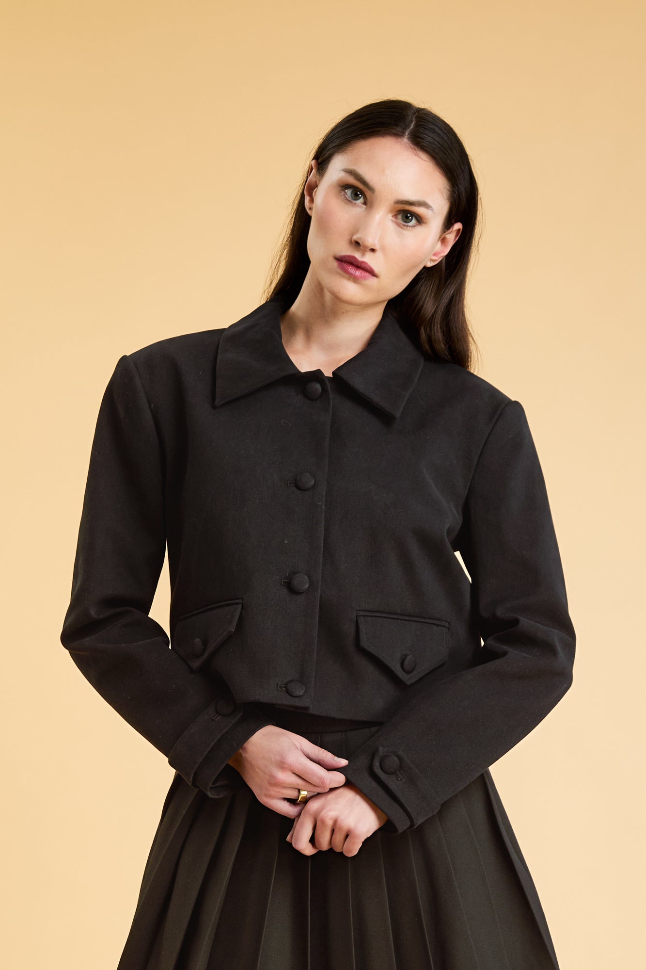 Woman wearing a Black Triplicity collared jacket and Black Triplicity pleated skirt on a beige background front close up