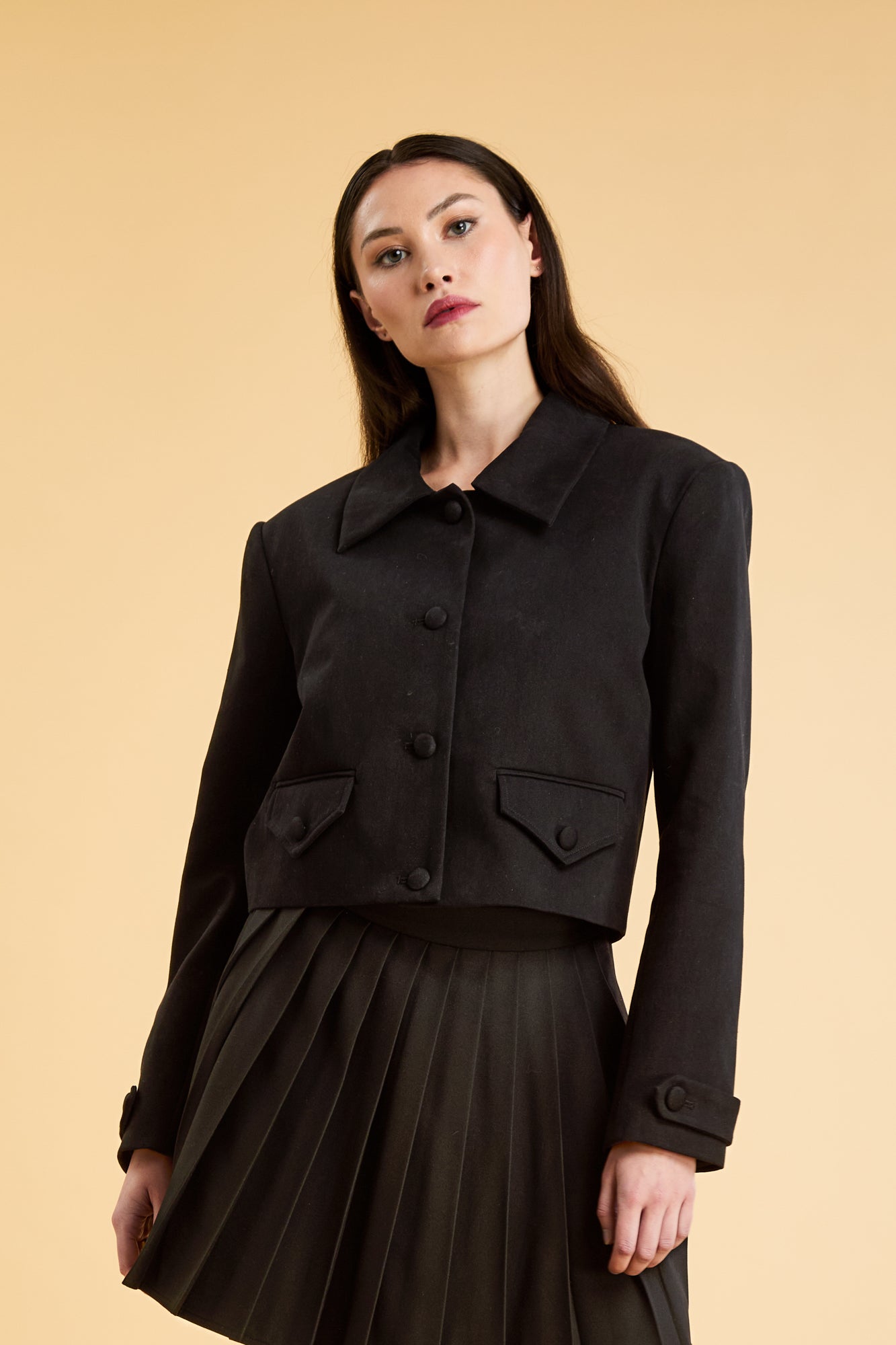 Woman wearing a Black Triplicity collared jacket and Black Triplicity pleated skirt on a beige background front close up