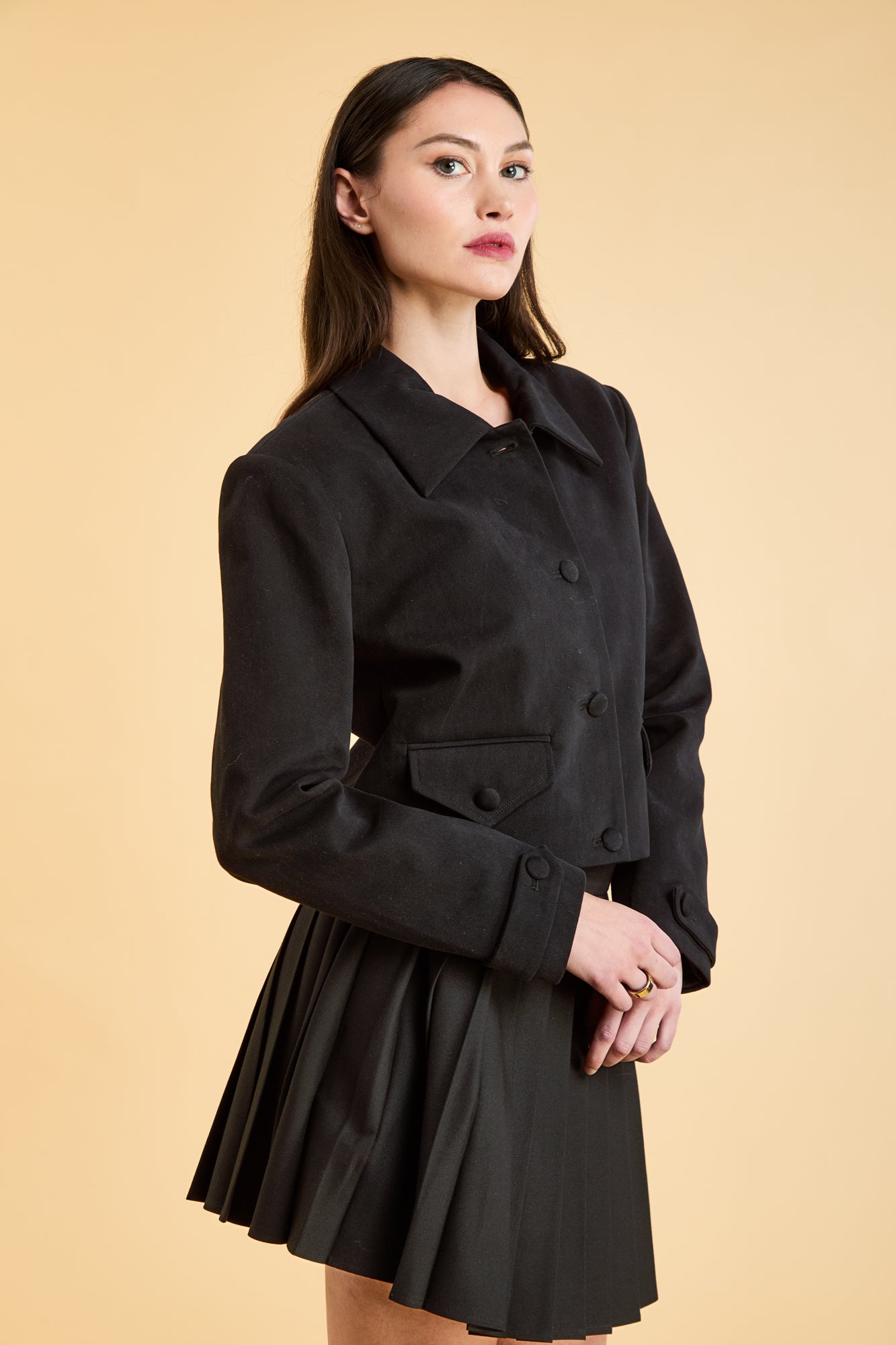 Woman wearing a Black Triplicity collared jacket and Black Triplicity pleated skirt on a beige background three quarter view close up