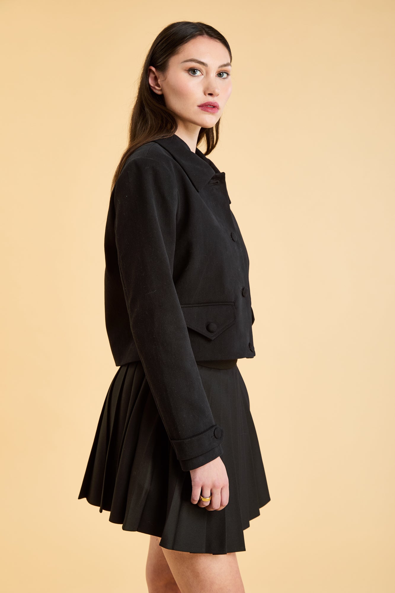 Woman wearing a Black Triplicity collared jacket and Black Triplicity pleated skirt on a beige background side close up