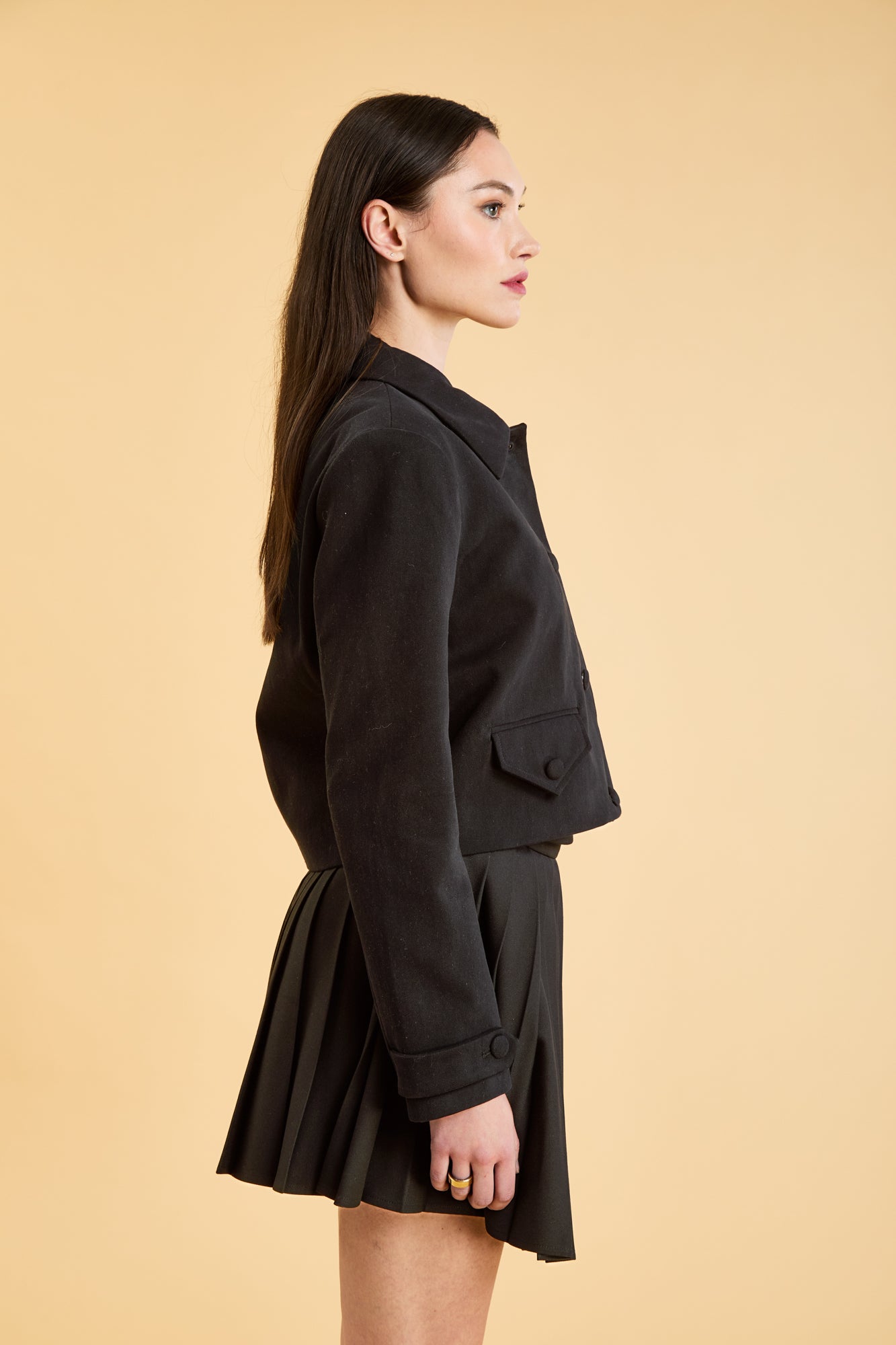 Woman wearing a Black Triplicity collared jacket and Black Triplicity pleated skirt on a beige background side close up