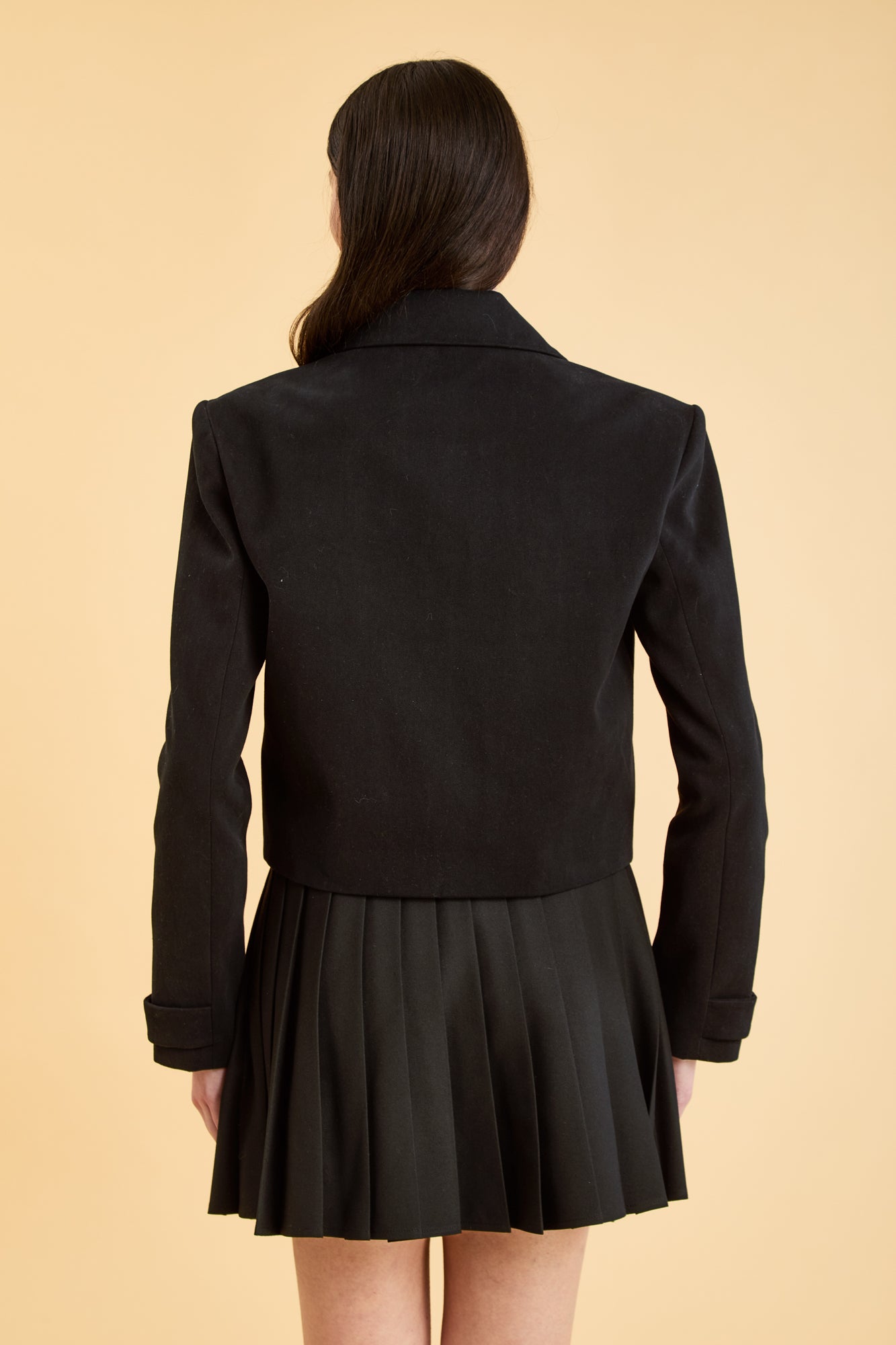 Woman wearing a Black Triplicity collared jacket and Black Triplicity pleated skirt on a beige background back close up