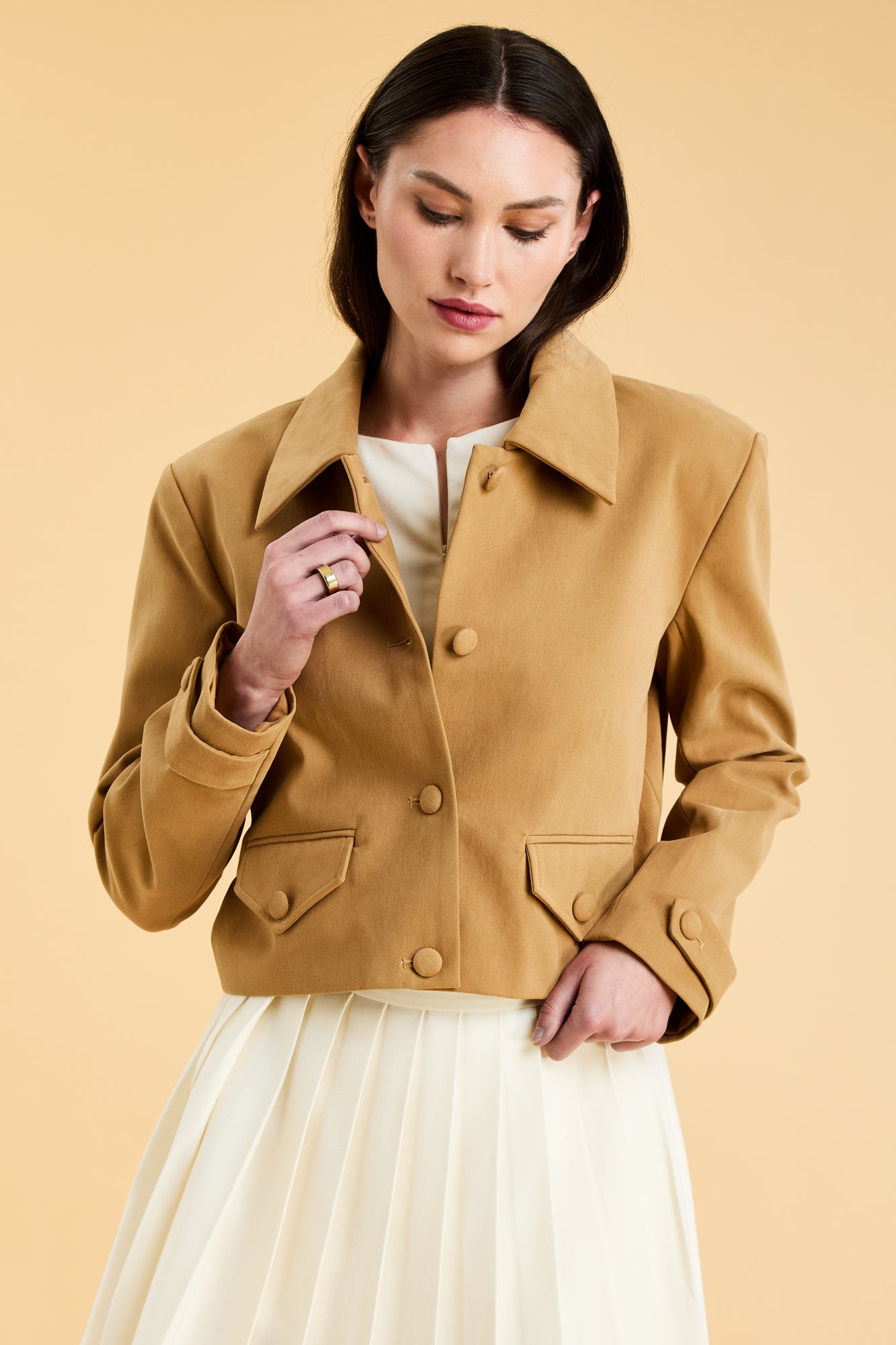 Woman wearing a Camel colored Triplicity collared tailored jacket on top of a White Triplicity pleated mini skirt on a beige background front view close up