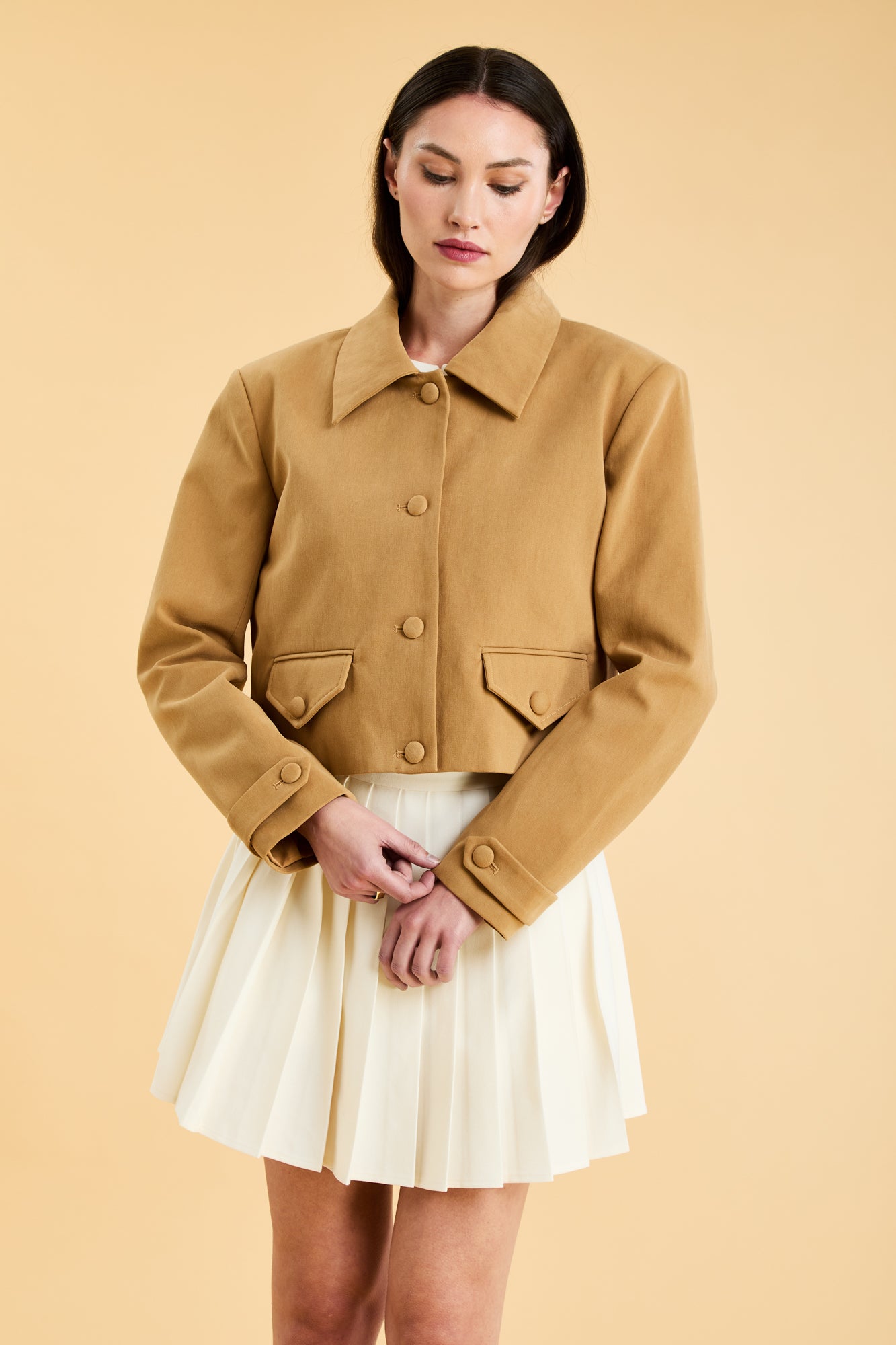 Woman wearing a Camel colored Triplicity collared tailored jacket on top of a White Triplicity pleated mini skirt on a beige background front view close up