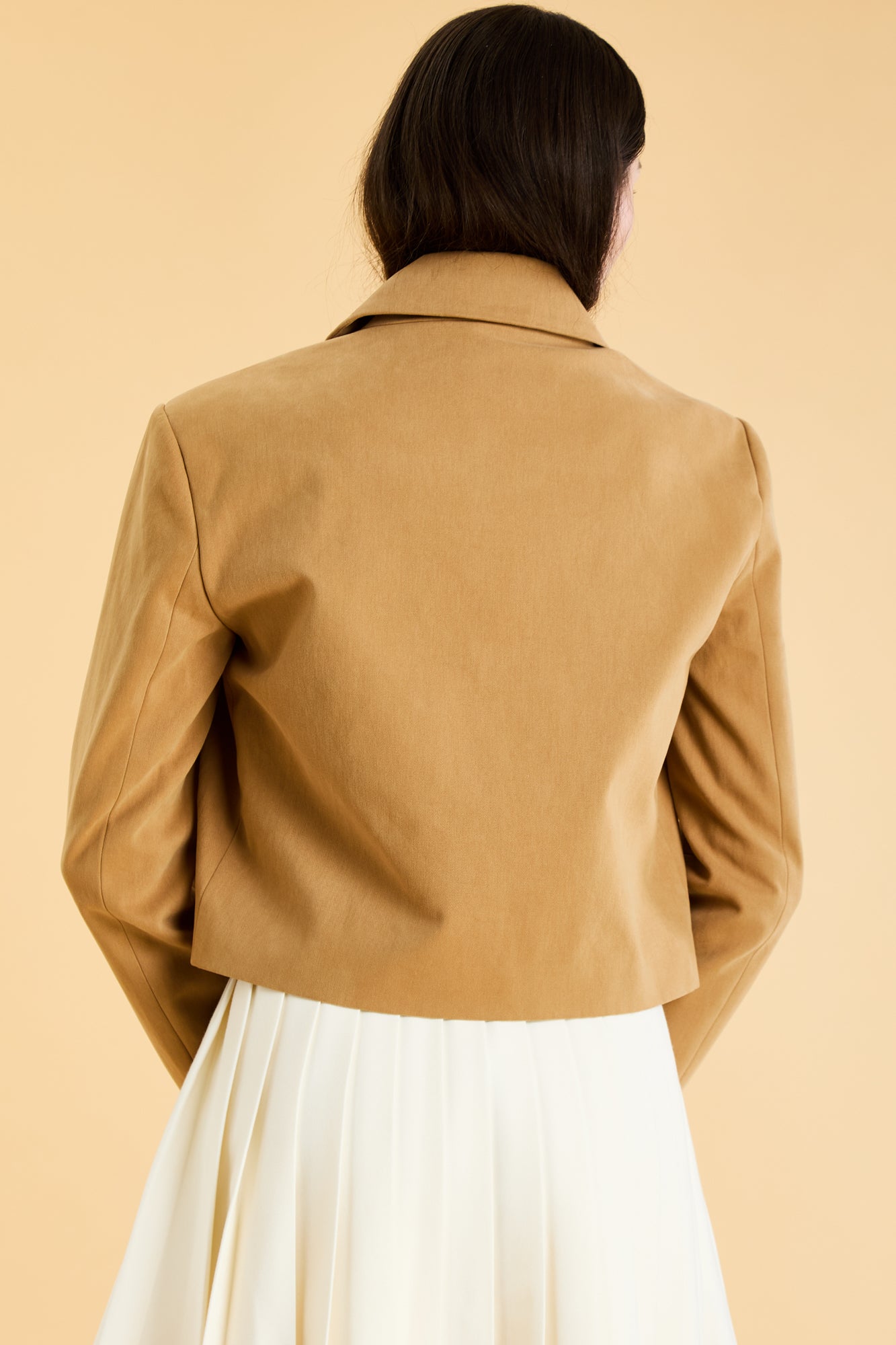 Woman wearing a Camel colored Triplicity collared tailored jacket on top of a White Triplicity pleated mini skirt on a beige background back view close up