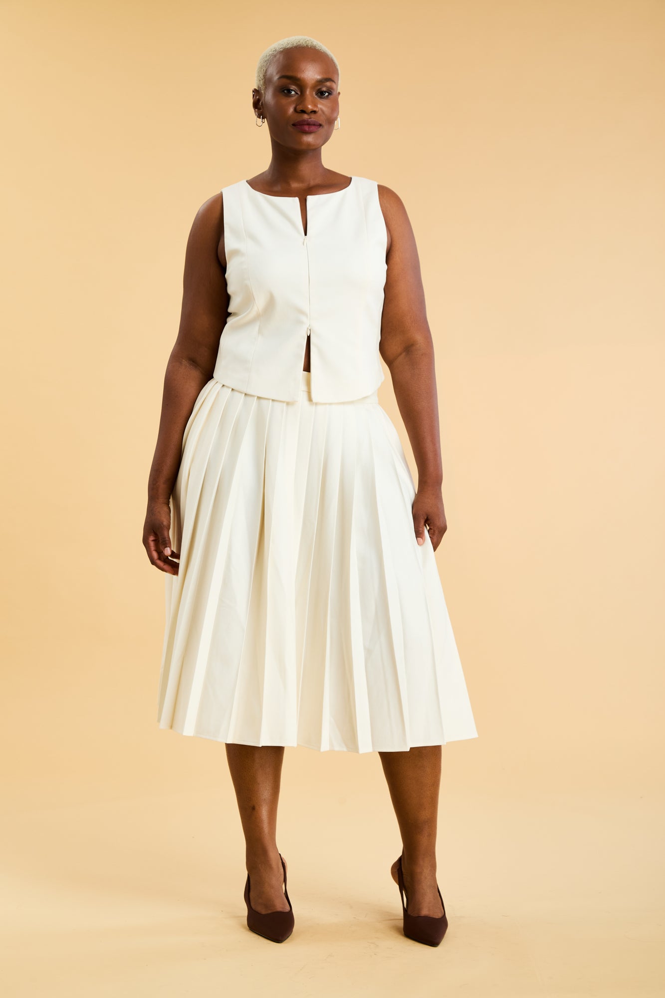 Woman wearing a White Triplicity double ended zipper sleeveless top and a White length White pleated skirt with a slit against a beige background full body front