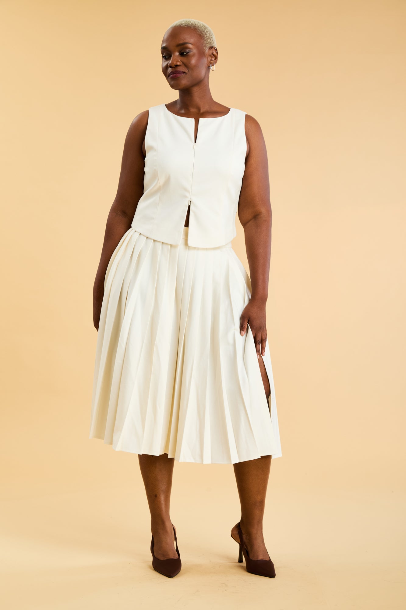Woman wearing a White Triplicity double ended zipper sleeveless top and a White length White pleated skirt with a slit against a beige background