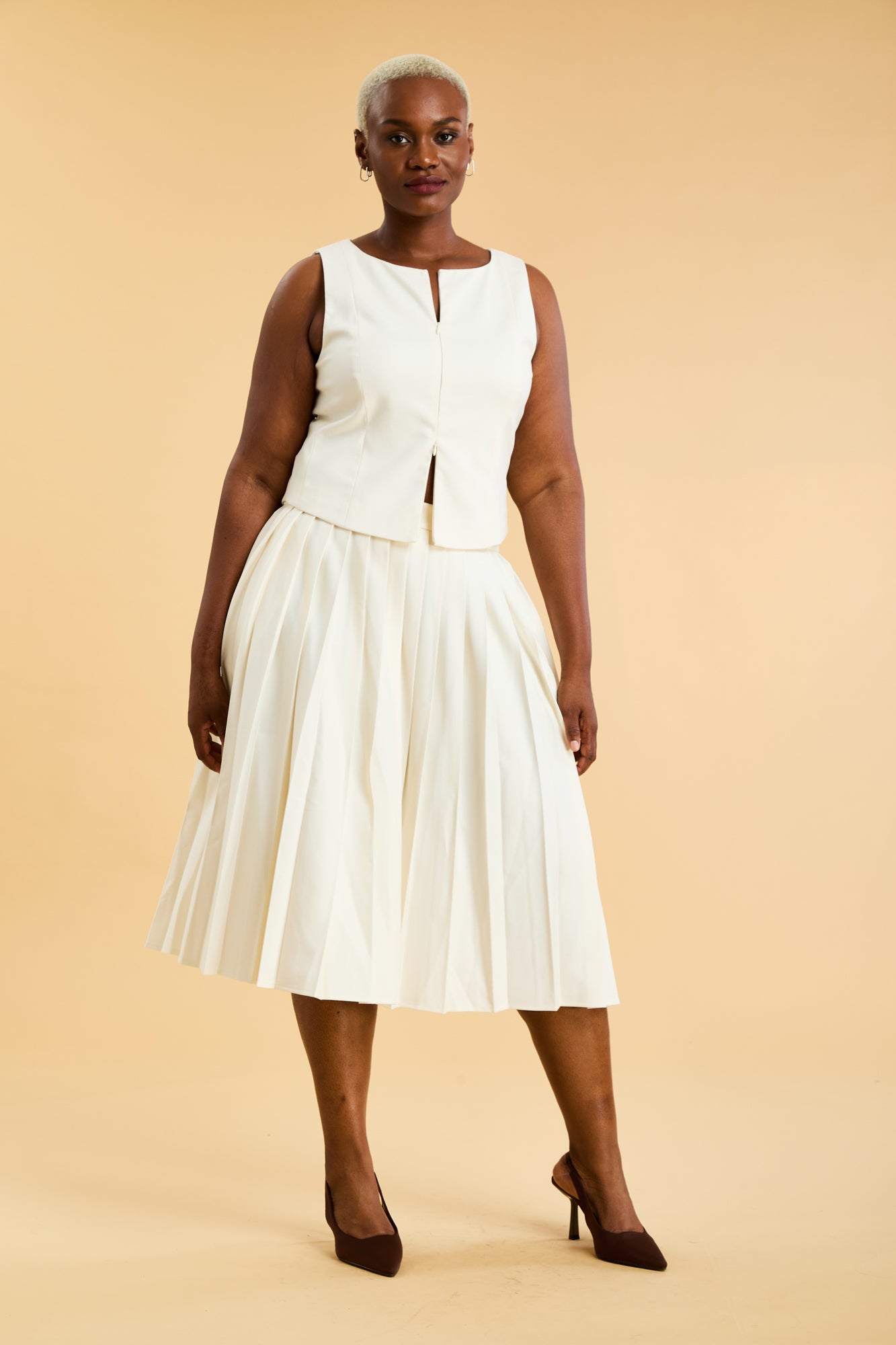 Woman wearing a White Triplicity double ended zipper sleeveless top and a White length White pleated skirt with a slit against a beige background