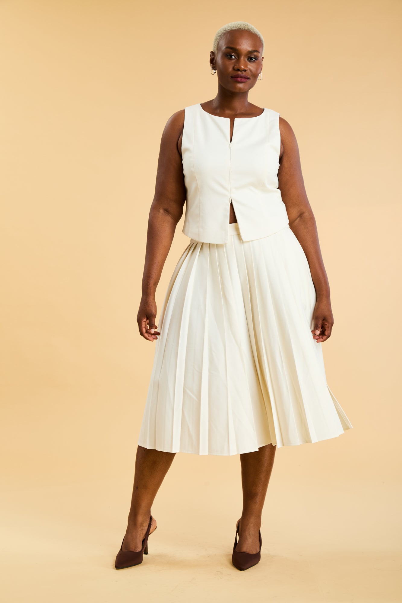 Woman wearing a White Triplicity double ended zipper sleeveless top and a White length White pleated skirt with a slit against a beige background