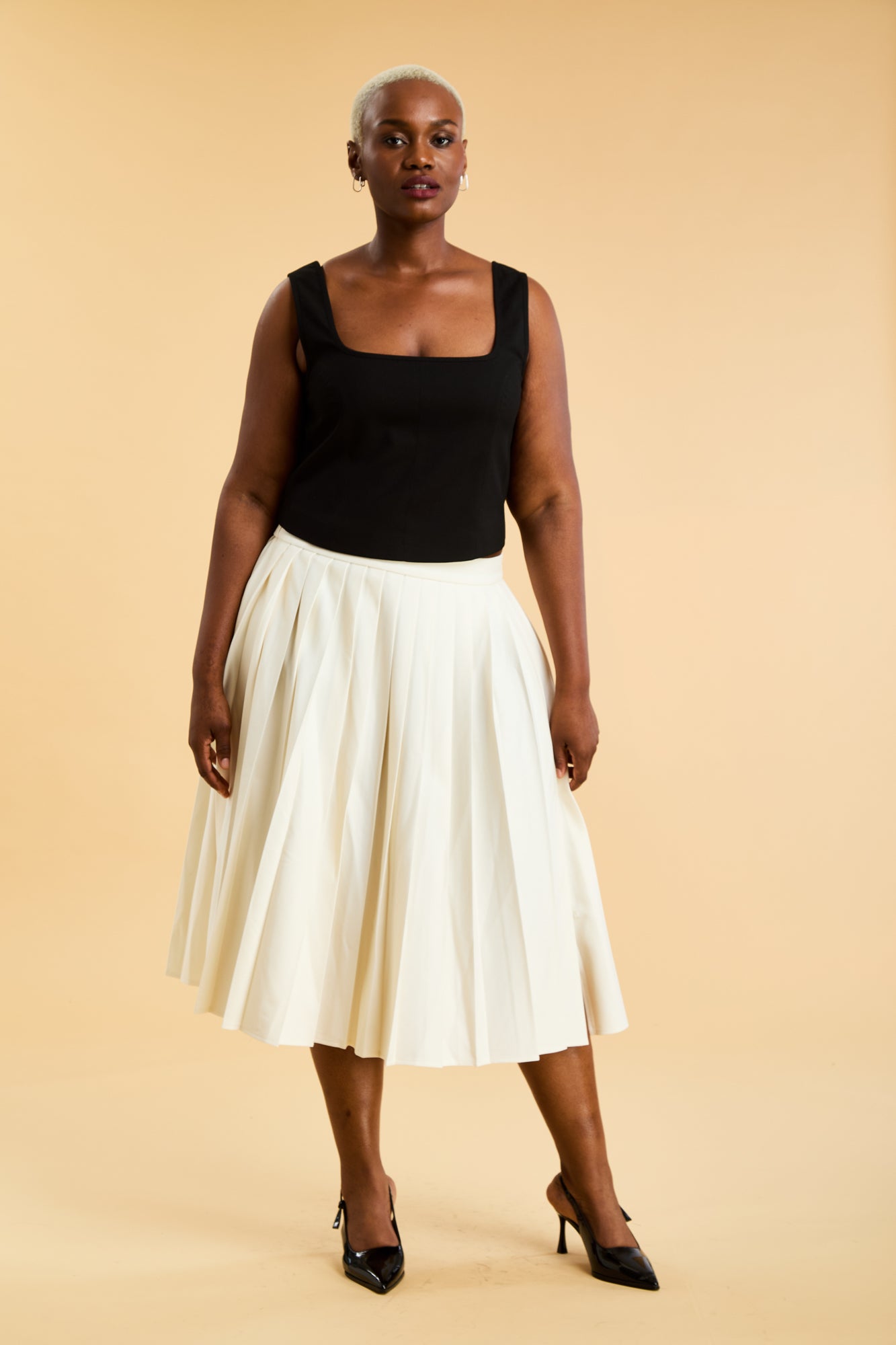 Woman wearing a Black Triplicity Eloise tank top and White Triplicity pleated skirt against a beige background front view full body