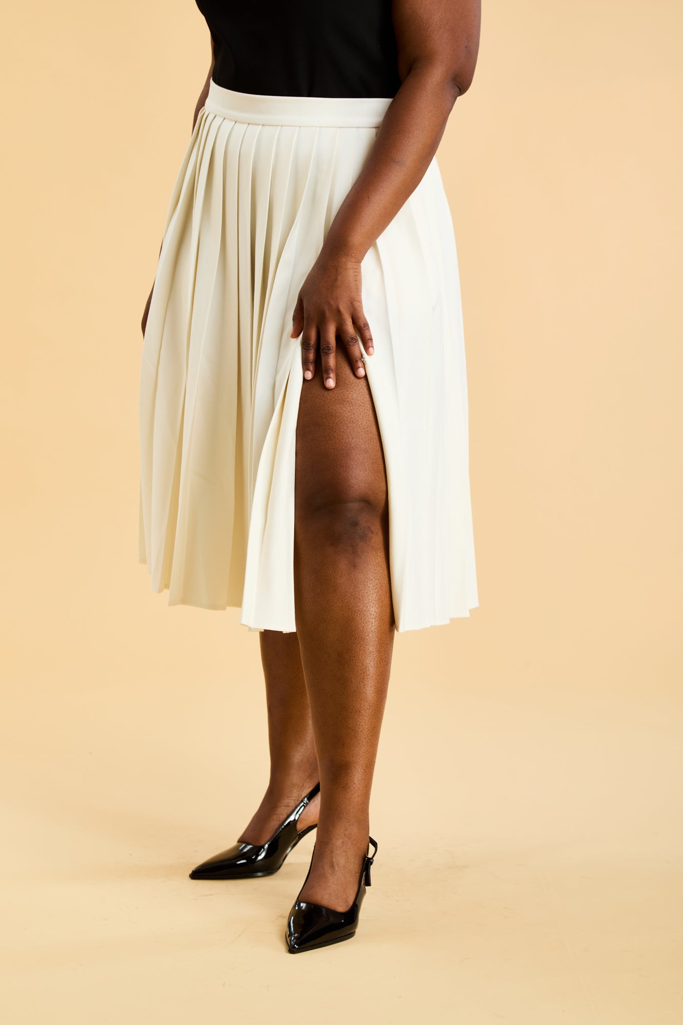 Bottom of a woman wearing a Black Triplicity sleeveless top and a White length White pleated skirt with a slit against a beige background close up