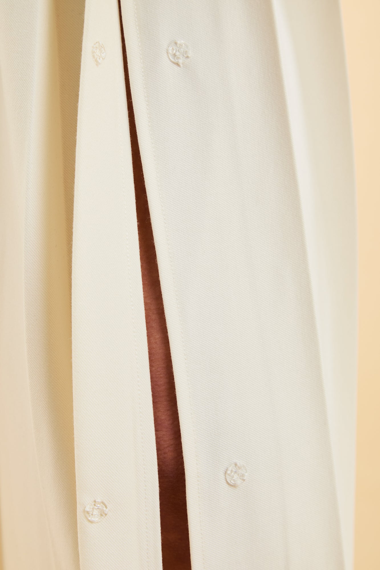 Close-up of a White Triplicity Pleated Midi skirt slit with a close up of two clear plastic snaps sewn into the sides of the slot on a beige background