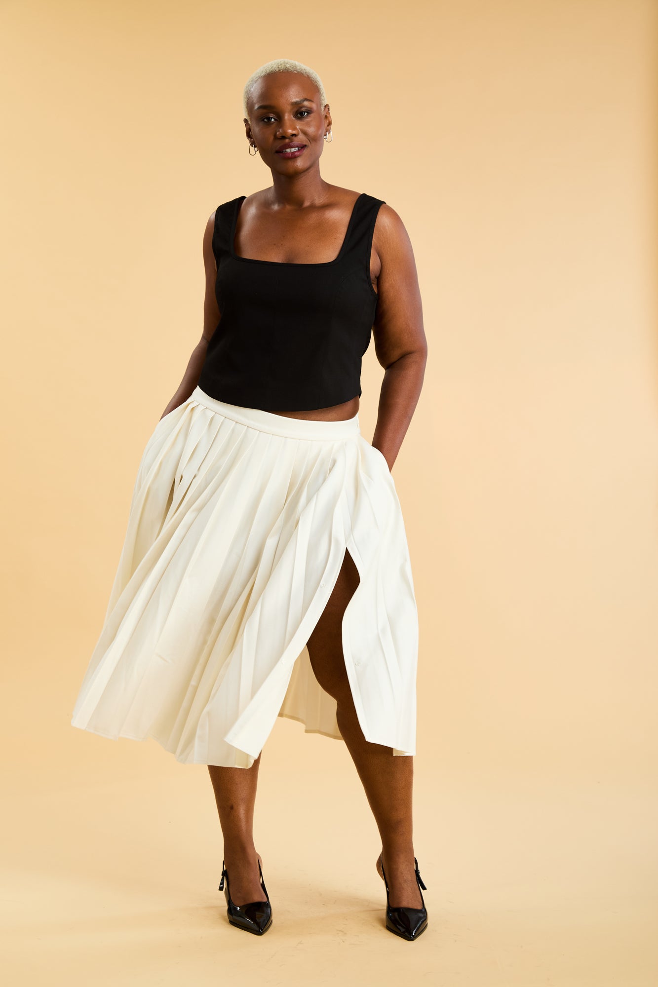 Woman wearing a Black Triplicity tank top and White pleated midi skirt with a slit against a beige background full body front view