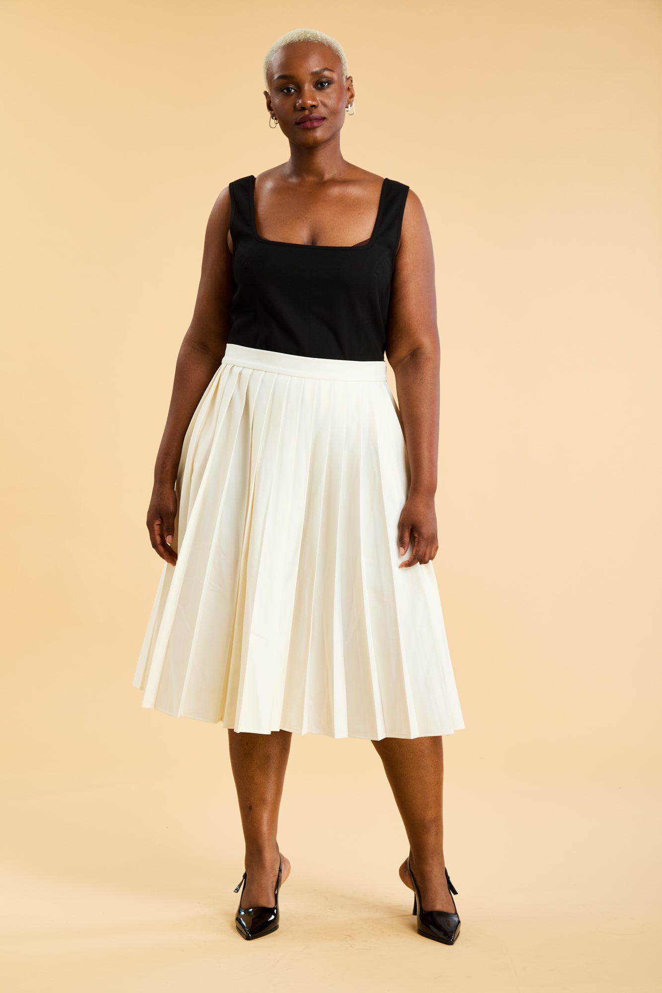 Woman wearing a Black Triplicity Eloise tank top and White Triplicity pleated skirt against a beige background front view full body