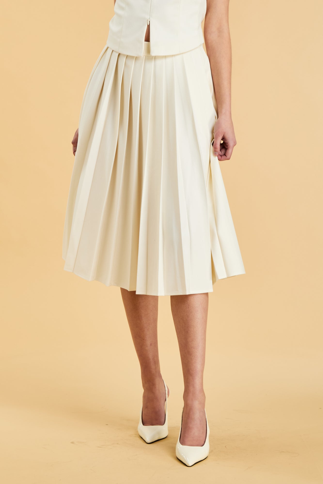 Close up of the bottom half of a woman wearing a White colored Triplicity Alice top and White Triplicity pleated midi skirt against a beige background front view