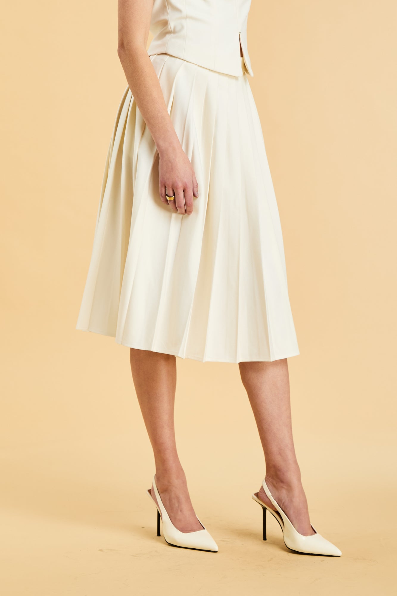Close up of the bottom half of a woman wearing a White colored Triplicity Alice top and White Triplicity pleated midi skirt against a beige background three quarter view
